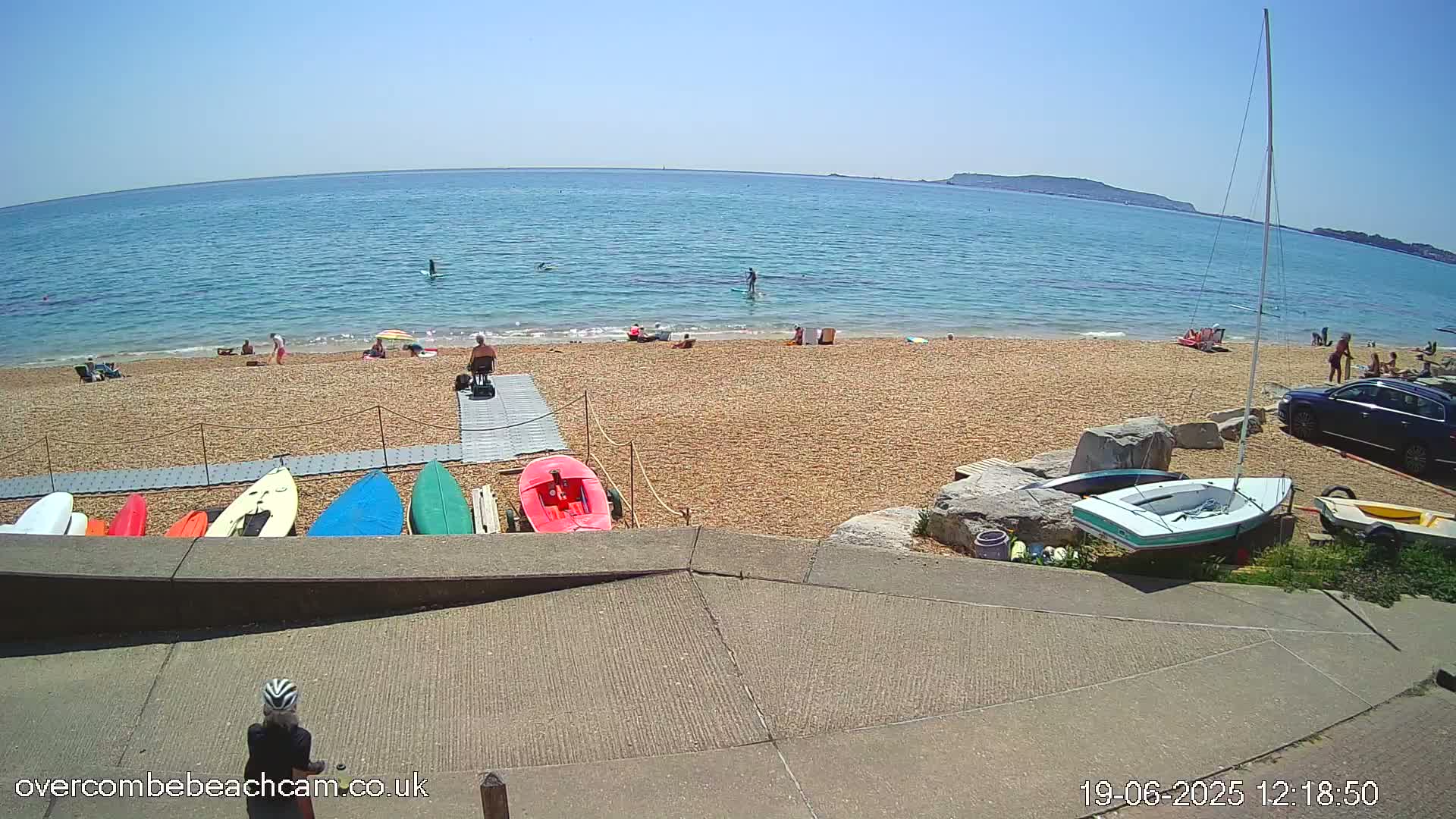 A sunny beach scene shows people relaxing and swimming in calm blue water, with several small boats and paddleboards along the shore.