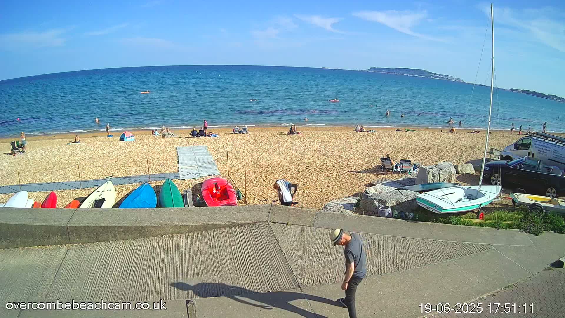 A sandy beach with scattered sunbathers, several small boats, and a man walking down a ramp, all under a sunny sky.