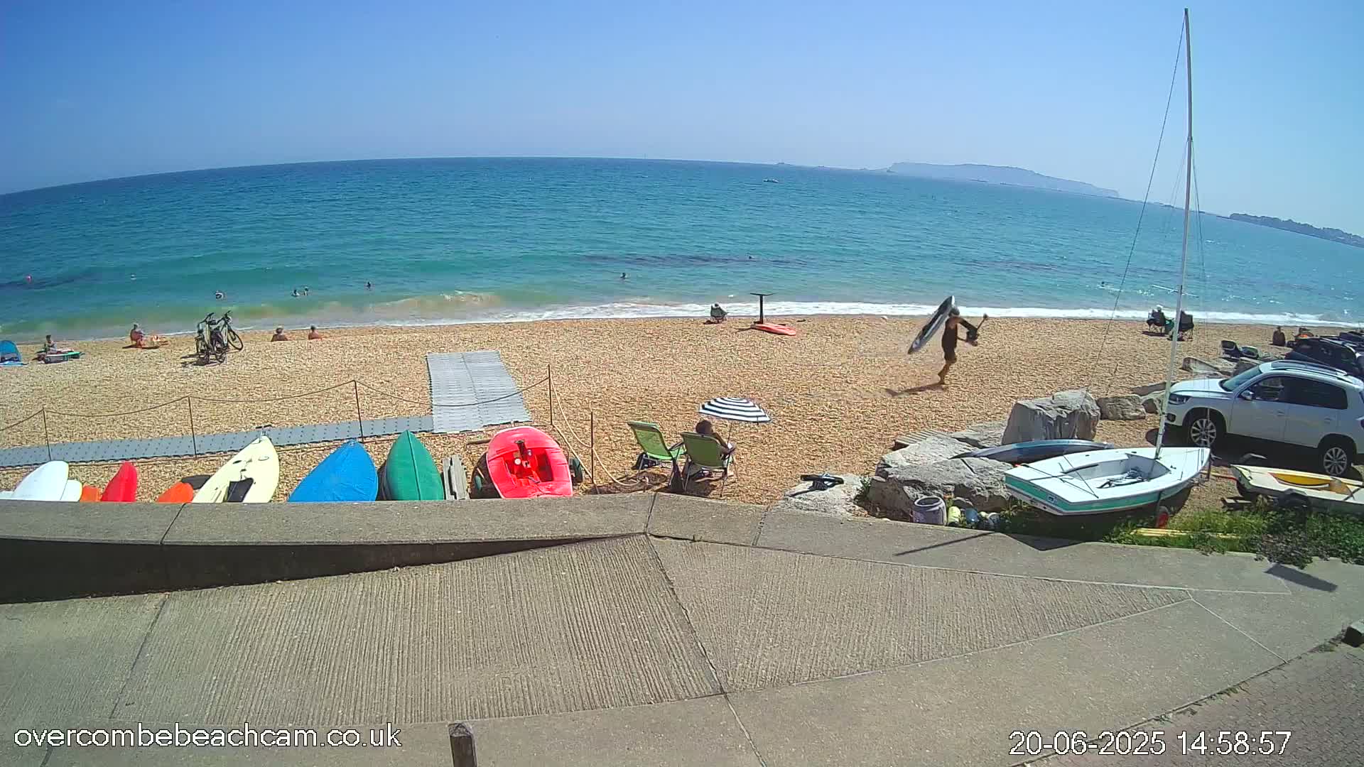 A sandy beach with a calm, blue ocean under a clear, sunny sky, shows several people relaxing and swimming, along with kayaks and a sailboat on the shore.