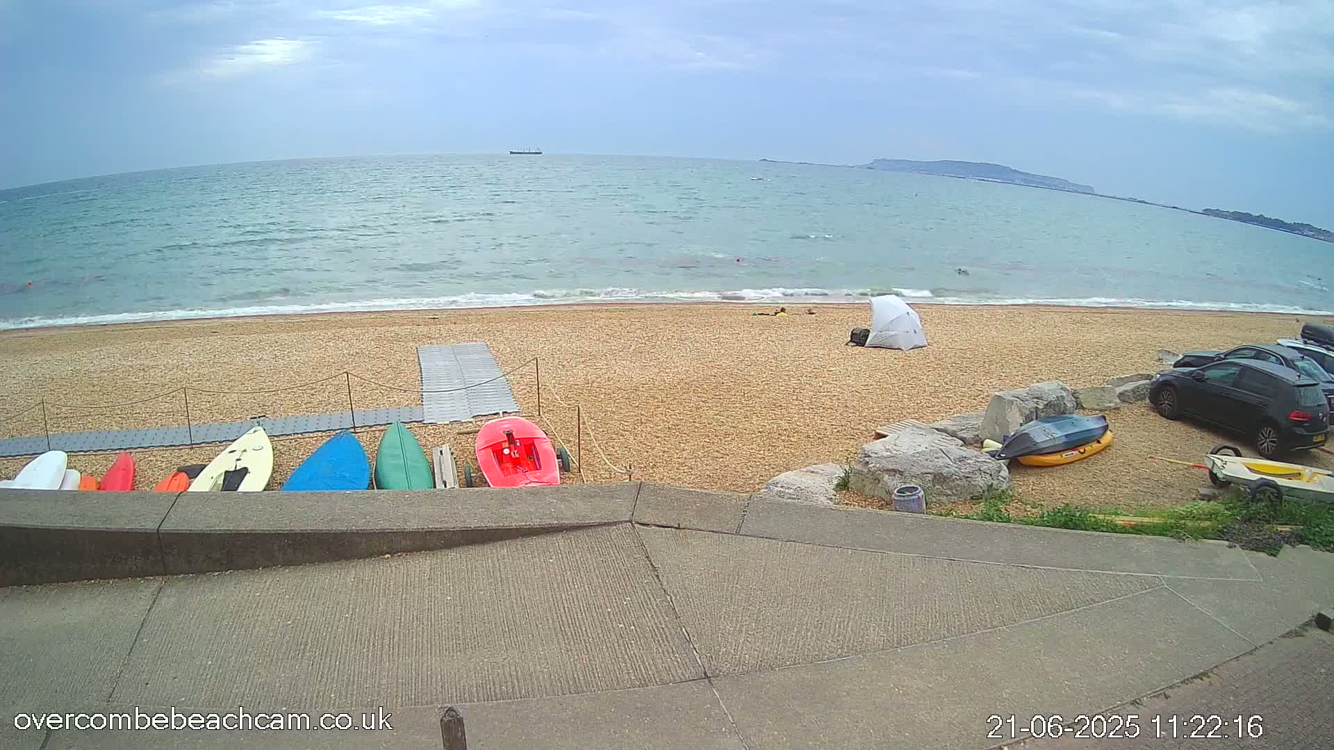 A sandy beach with several small boats and kayaks lined up near a paved walkway, with a few people relaxing on the beach under an overcast sky and calm sea in the background.