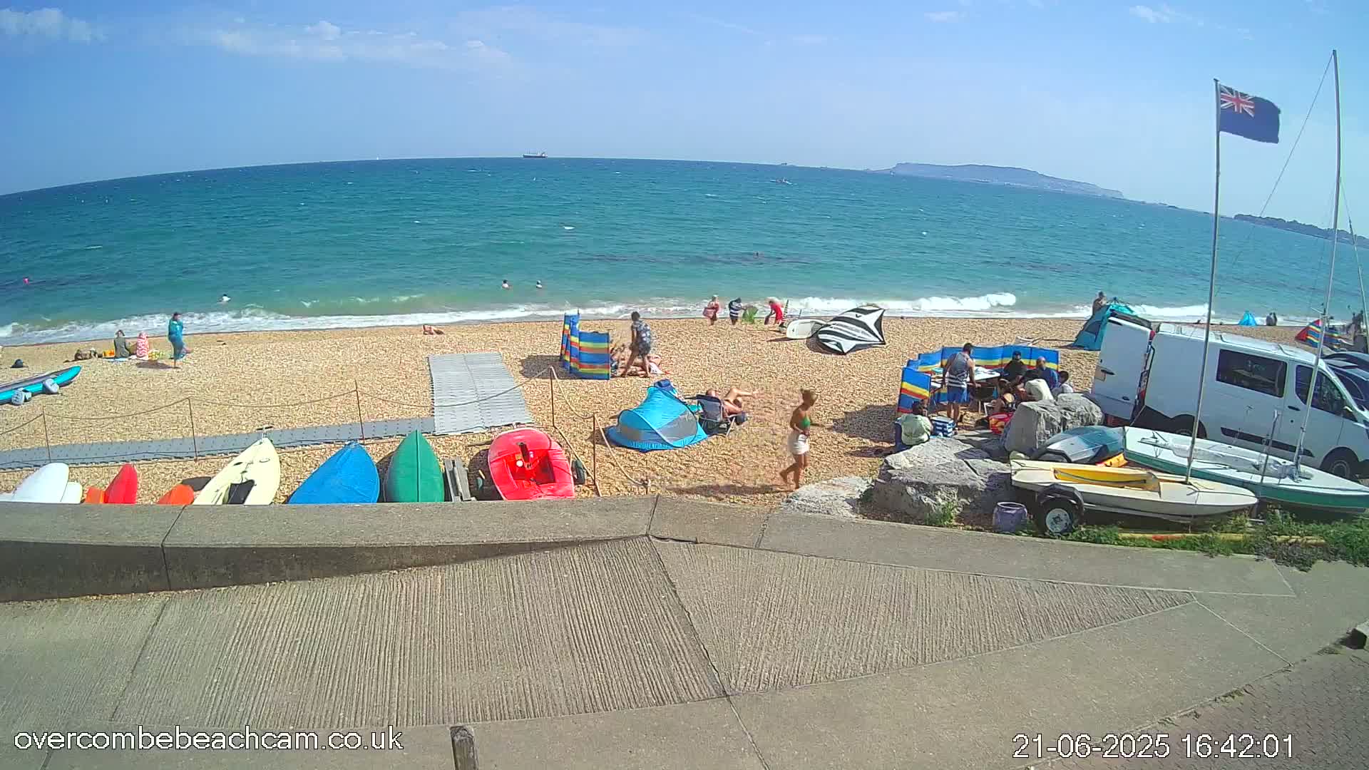 A sunny beach scene shows people relaxing and swimming in a turquoise ocean, with kayaks and small sailboats on the sand near a van.