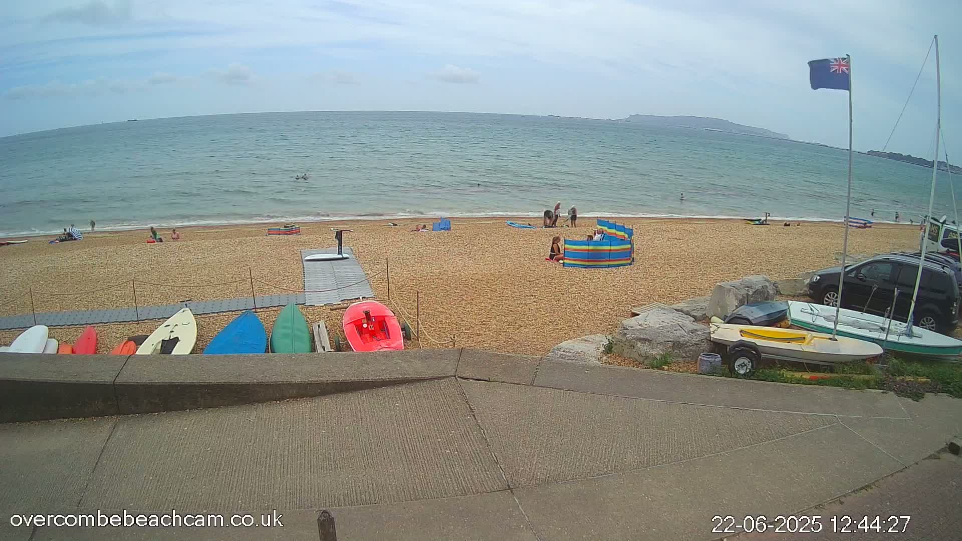 A mostly sunny day at a pebble beach shows several people relaxing and swimming in the ocean, with various watercraft and windbreaks on the shore, and several parked cars nearby.