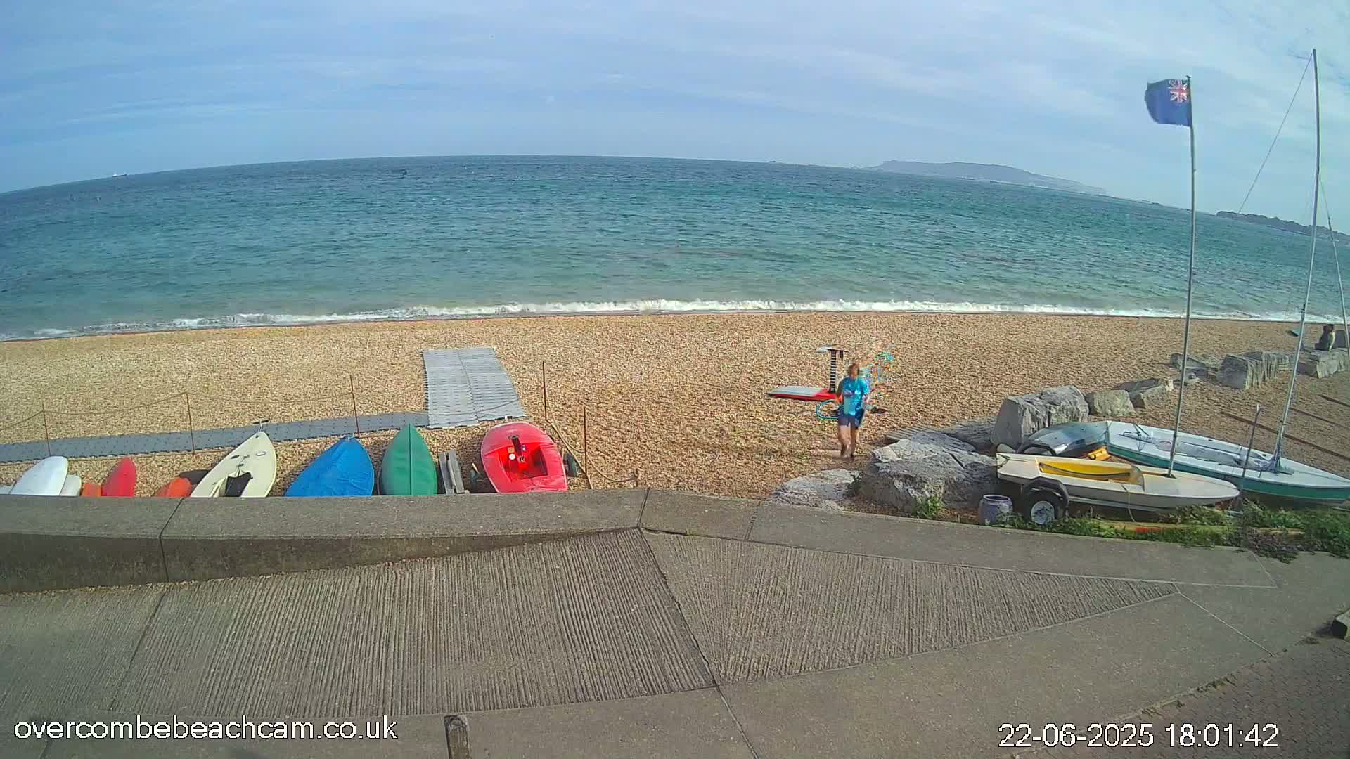 A person stands on a pebble beach next to several small boats and kayaks near a calm ocean under a clear sky.