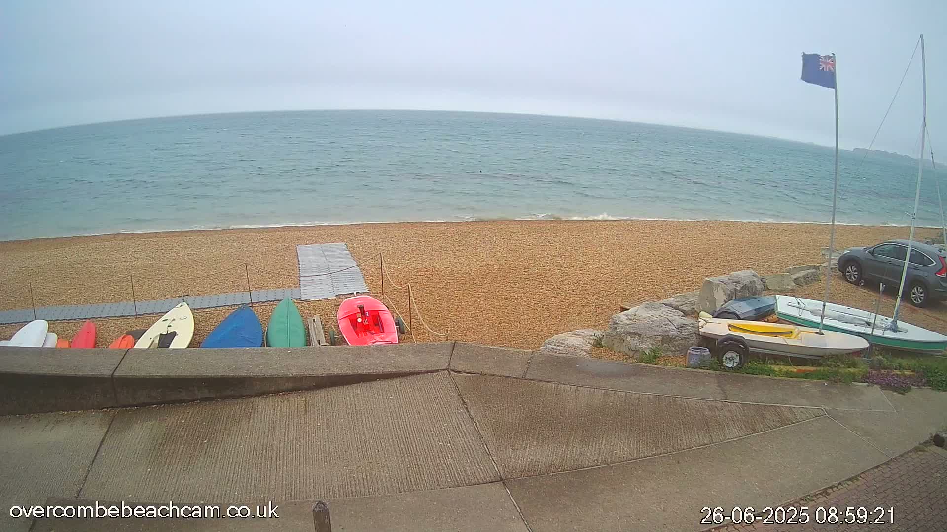 Several small boats and kayaks are stored on a pebble beach next to a calm, gray sea under an overcast sky.