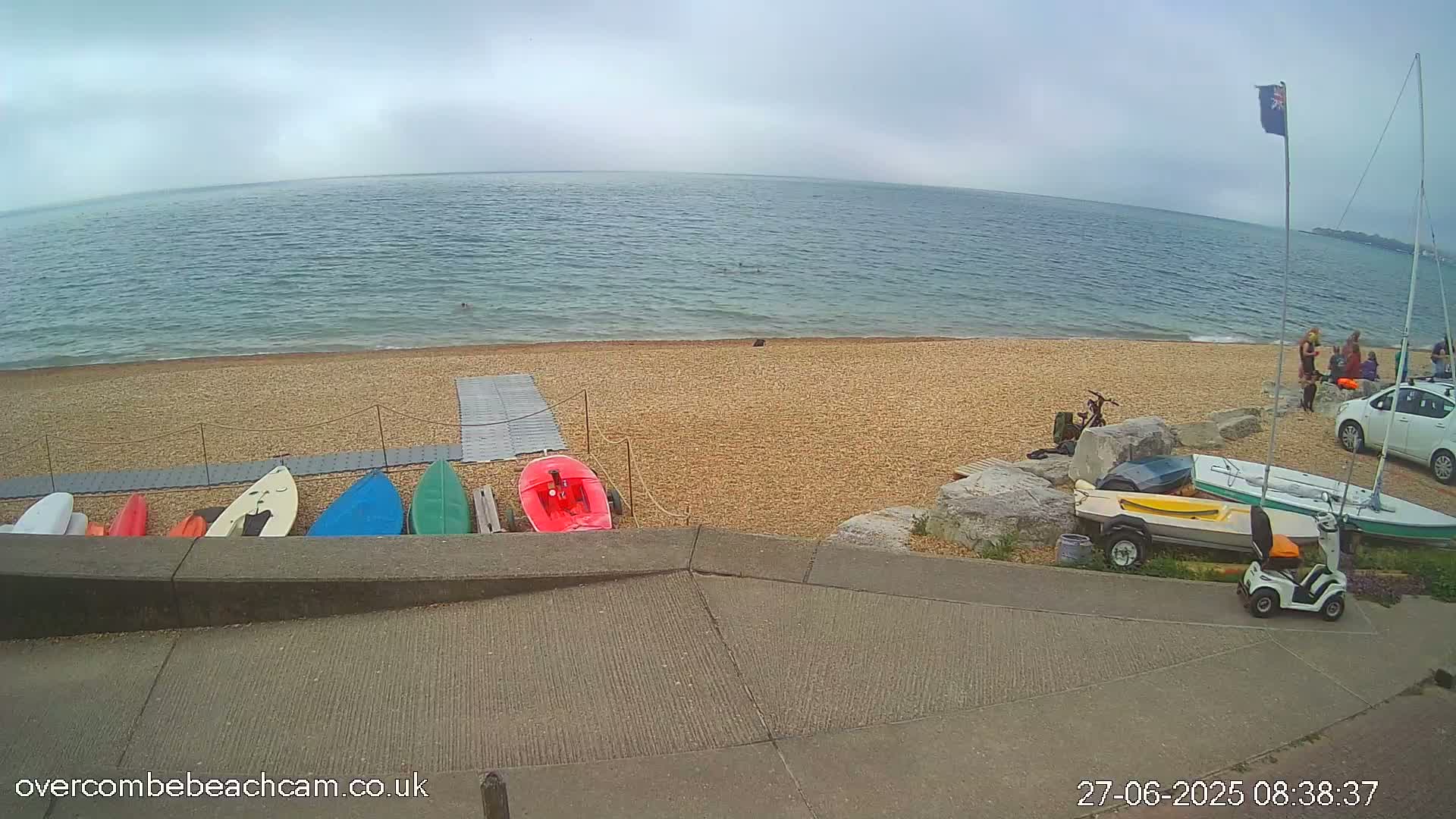A pebble beach with several small boats and a mobility scooter parked near a calm sea under an overcast sky.