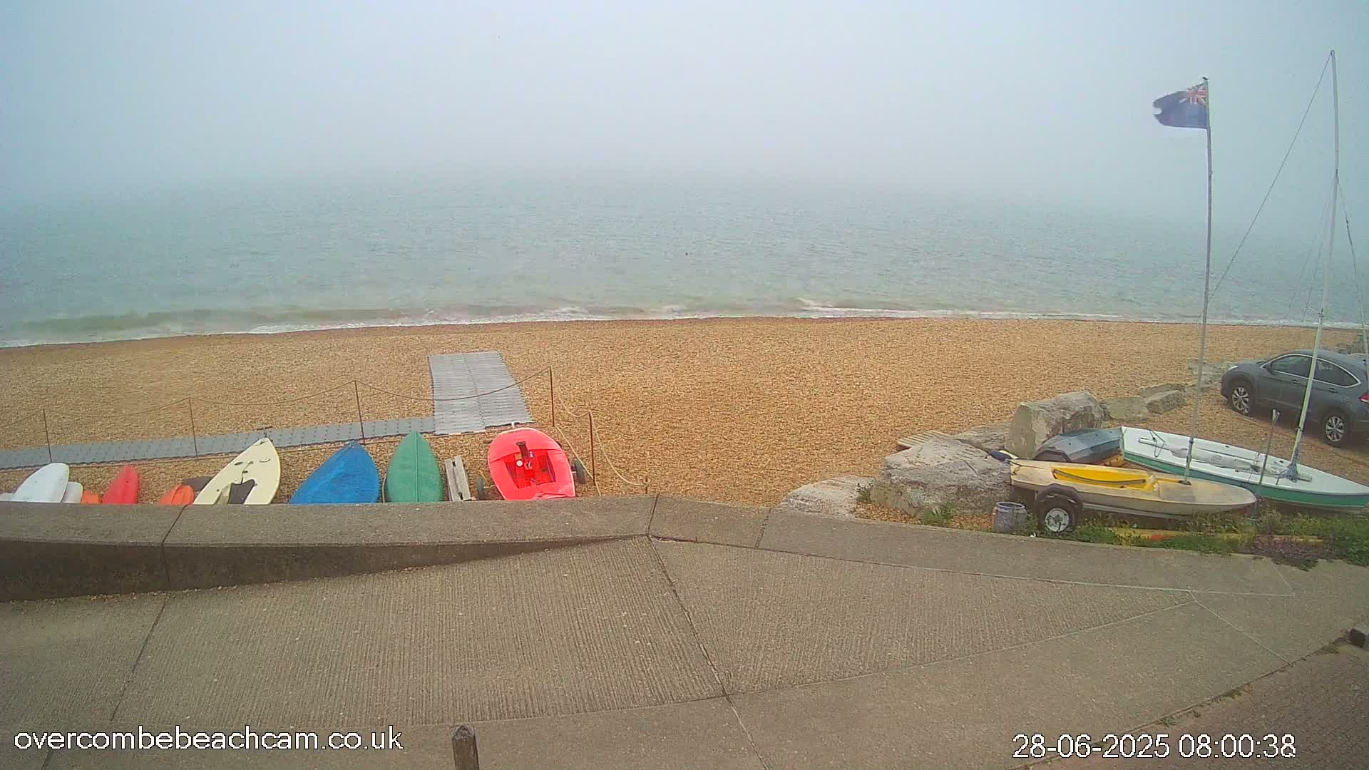 A foggy day at a pebble beach shows several small boats and kayaks stored on a ramp, with a sailboat and car parked nearby.