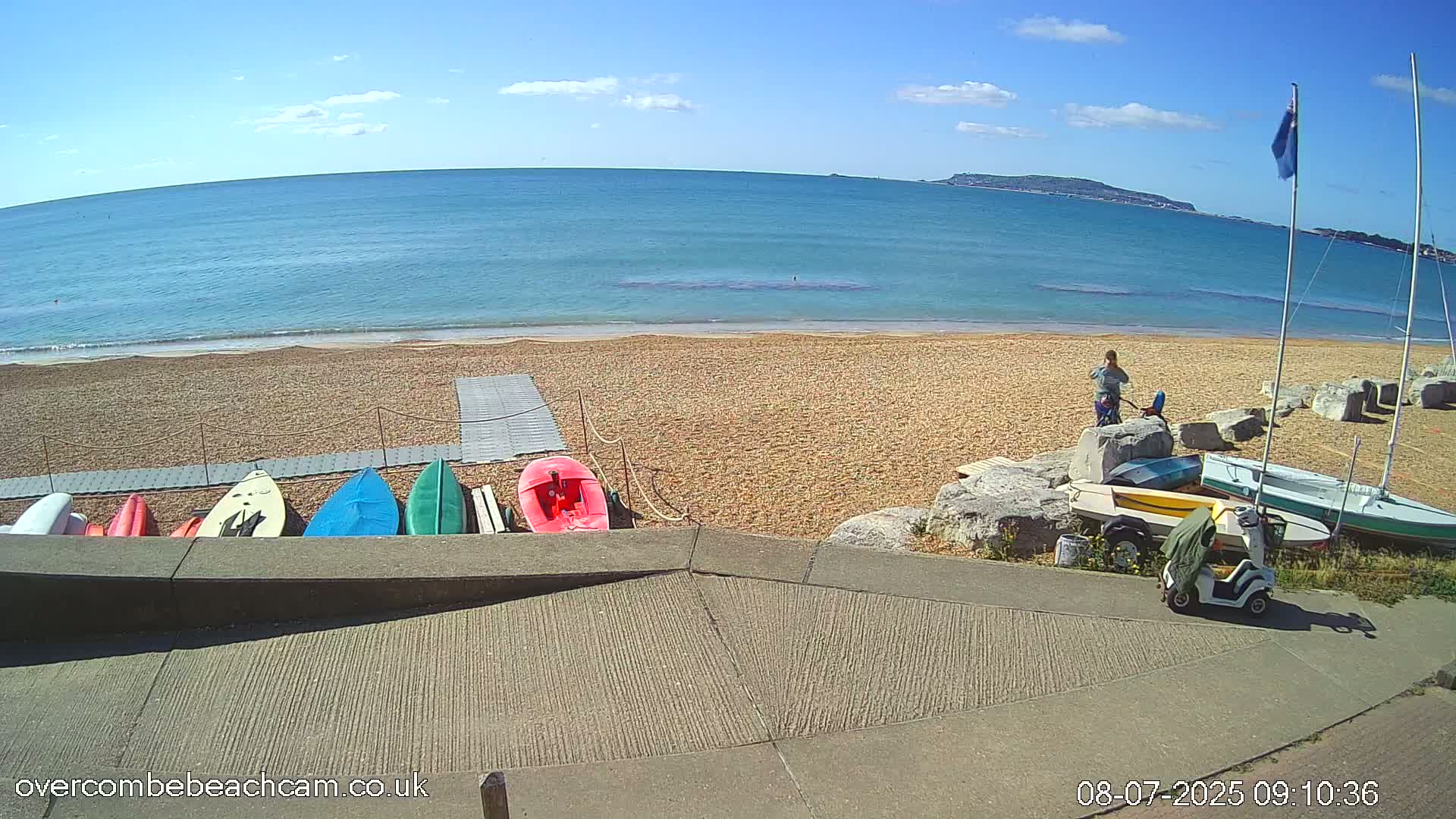 A sandy beach with several kayaks and sailboats near a paved area, overlooks a calm ocean under a mostly sunny sky.