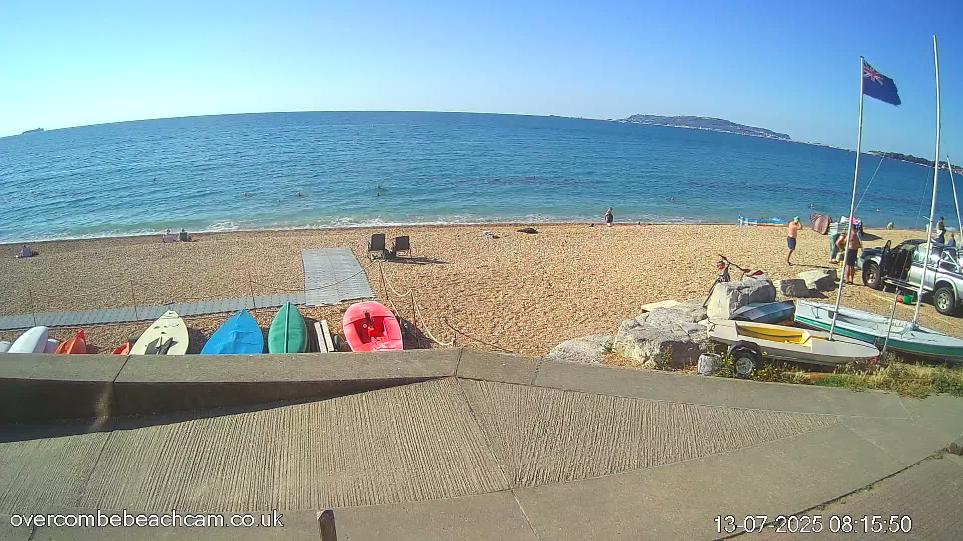 A sunny beach scene shows a calm blue ocean, a pebbly shore with several small boats and kayaks, and a few people enjoying the water and sun.