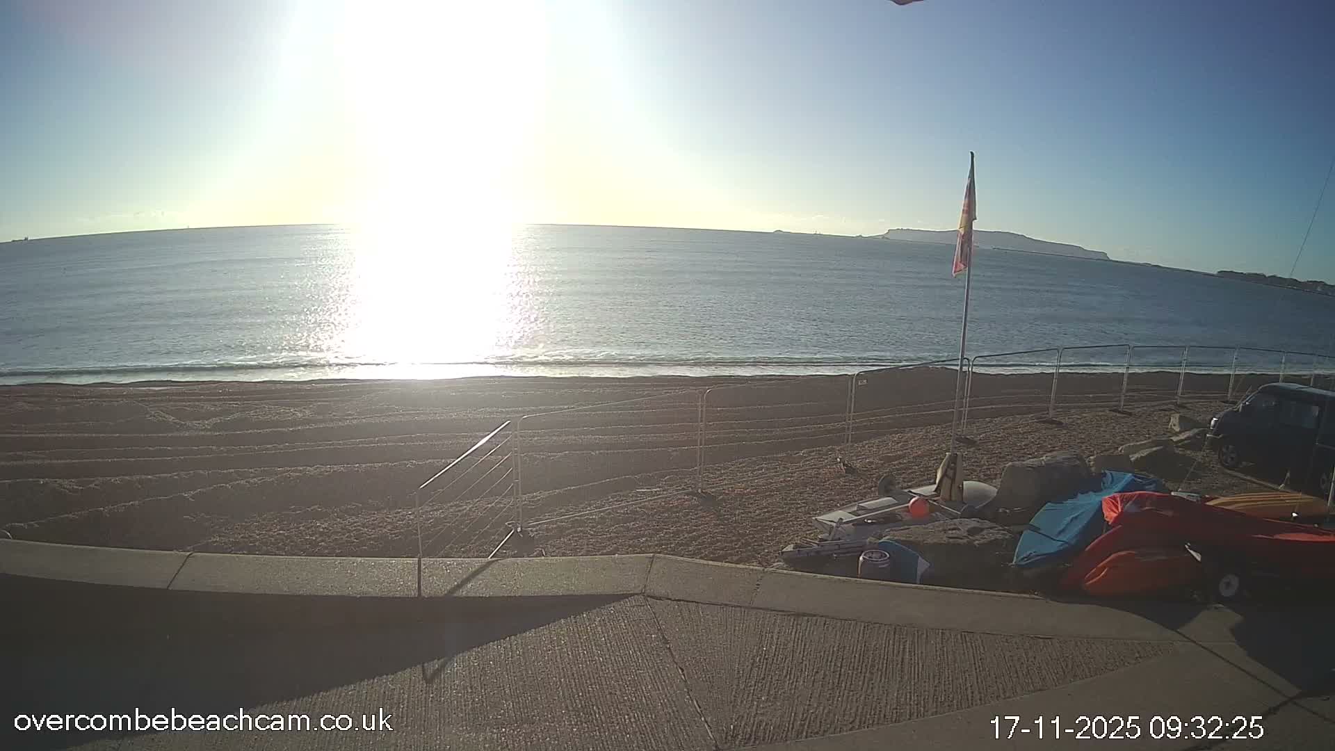 A bright, sunny day with a clear blue sky illuminates a shingle beach featuring gentle waves, a flag, several small boats and kayaks, and distant land across the calm bay.
