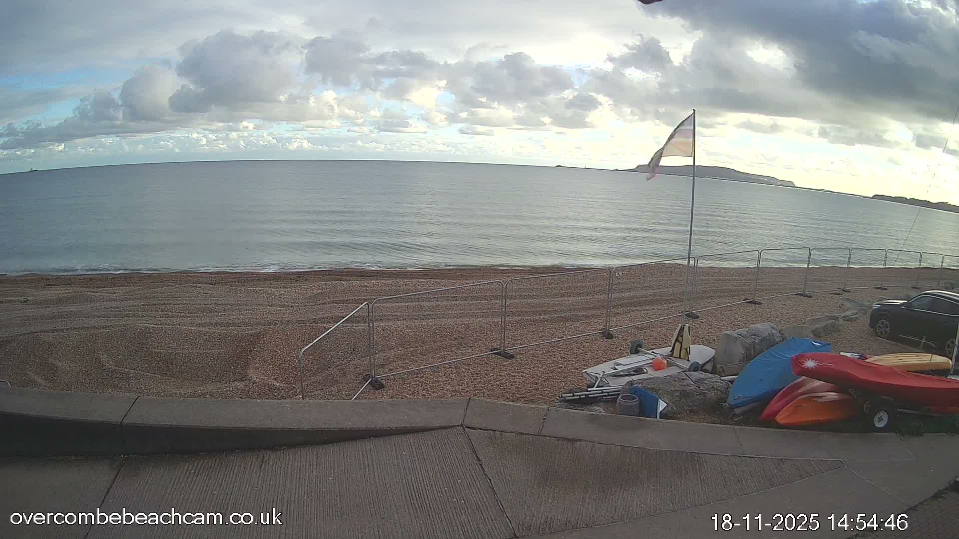 A bright, sunny day with a clear blue sky illuminates a shingle beach featuring gentle waves, a flag, several small boats and kayaks, and distant land across the calm bay.