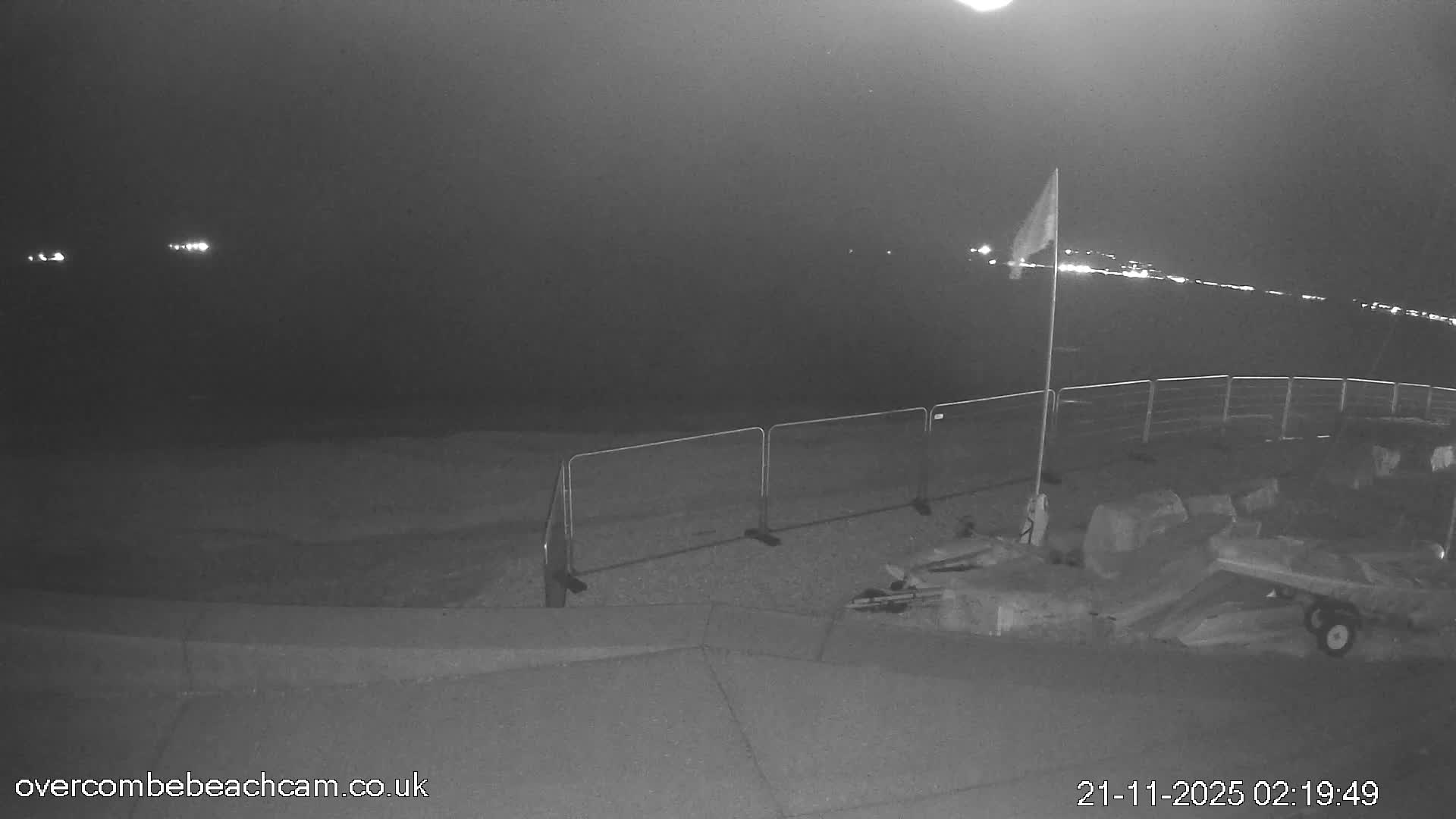 A bright, sunny day with a clear blue sky illuminates a shingle beach featuring gentle waves, a flag, several small boats and kayaks, and distant land across the calm bay.