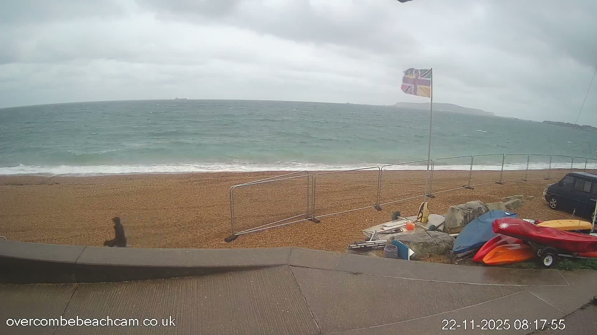 A person walks along a pebbly beach with choppy turquoise-grey seas under an overcast sky, while a Union Jack flag flies in the wind and various kayaks and beach equipment are stored near a van on the right.