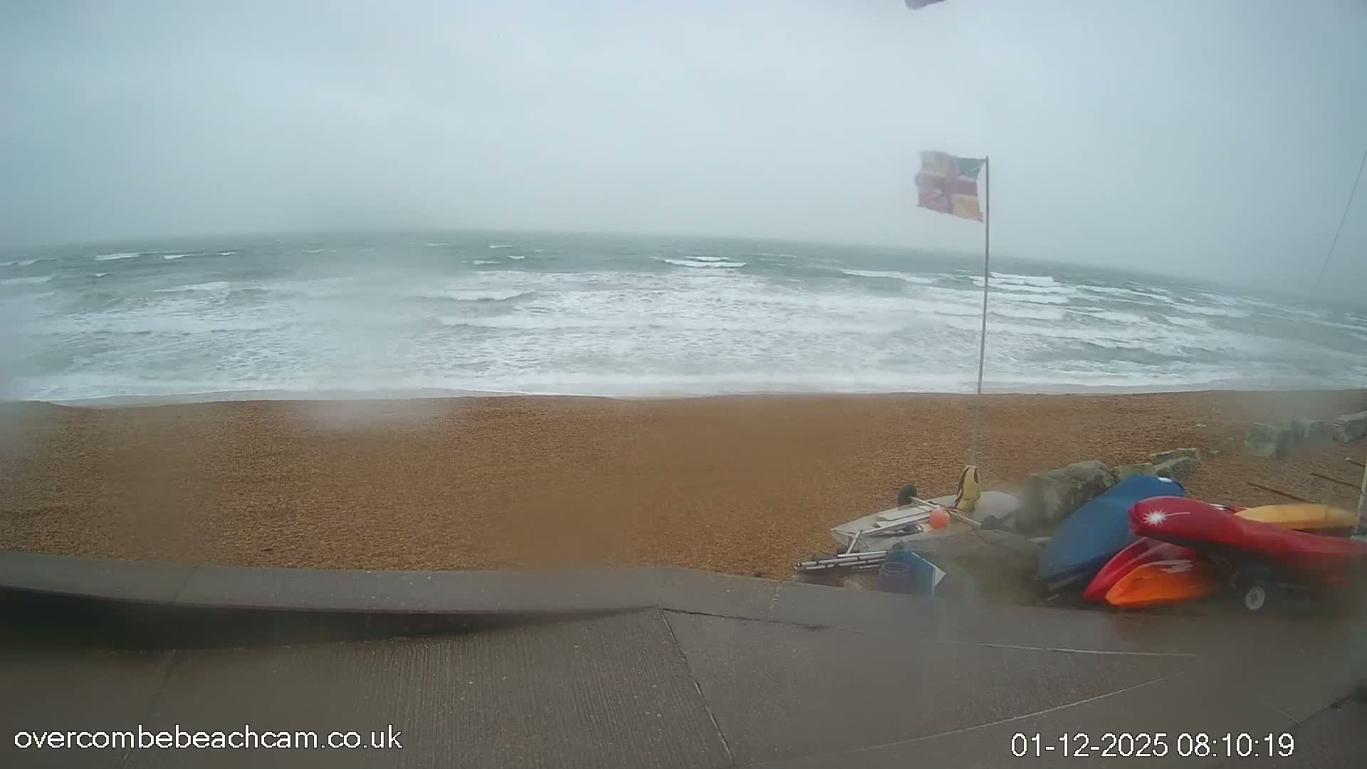 A blustery day at a pebble beach features rough, white-capped waves crashing onto the shore, a flag whipping in the wind, and several kayaks and small boats stored near a concrete promenade under an overcast sky.