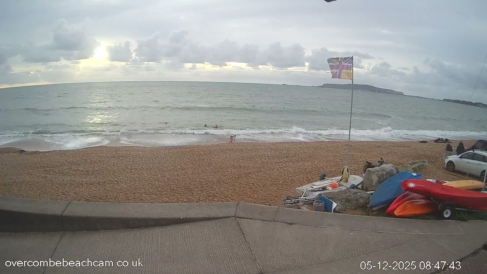 On an overcast day with the sun peeking through, people swim in the choppy sea at a shingle beach where a Union Jack flag flies and various boats and kayaks are stored near a parked car.