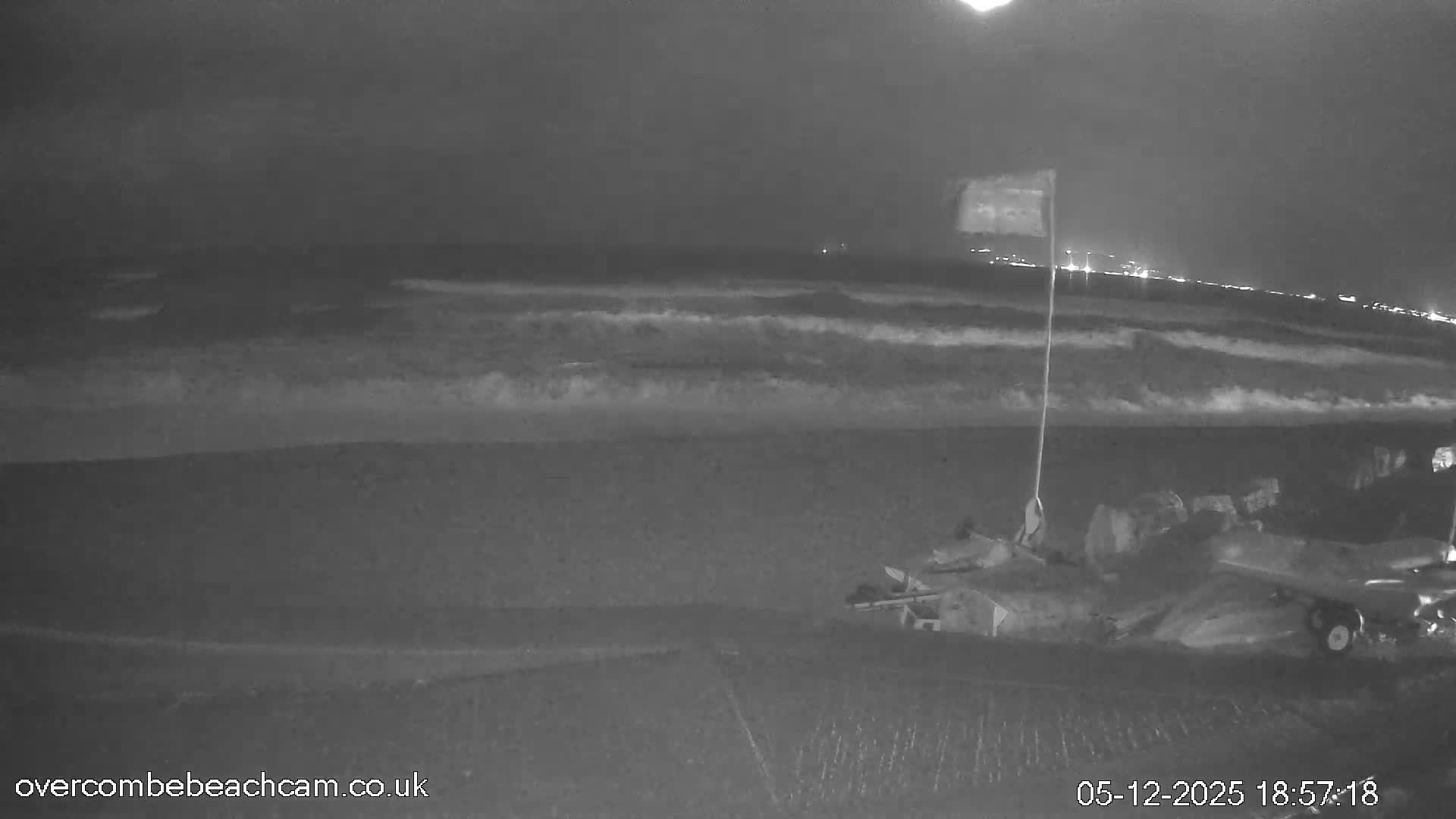 A dark, cloudy night scene at a beach shows waves breaking on the shore under a full moon, with distant city lights illuminating the horizon and some beach equipment and a flag visible in the foreground.