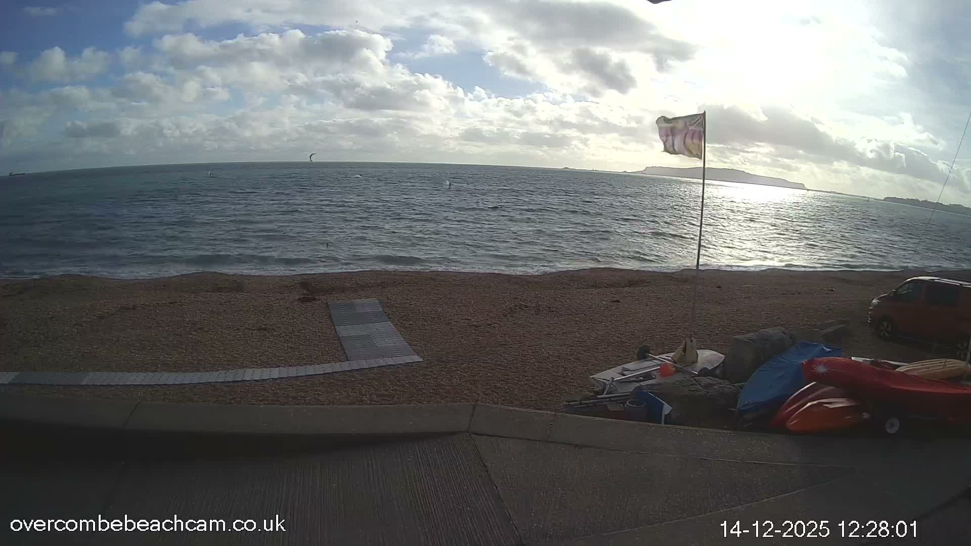 A dark, cloudy night scene at a beach shows waves breaking on the shore under a full moon, with distant city lights illuminating the horizon and some beach equipment and a flag visible in the foreground.
