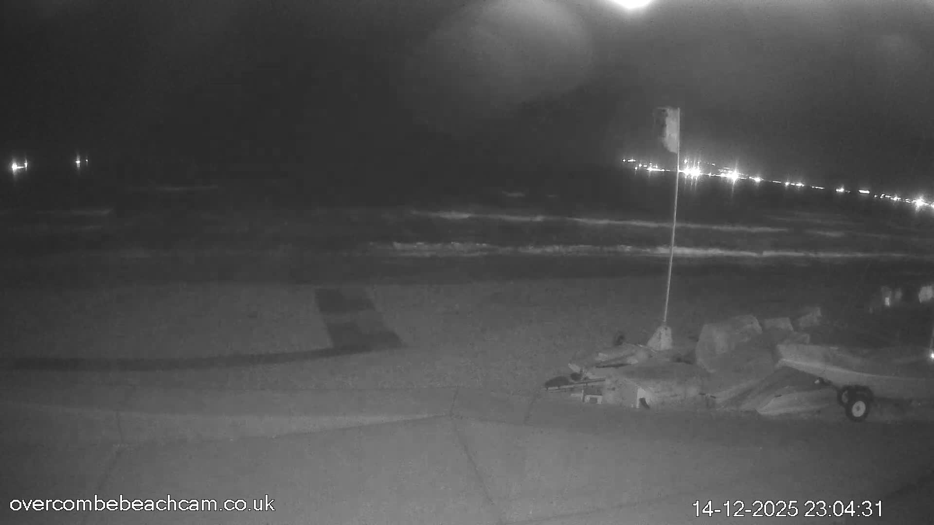 A dark, cloudy night scene at a beach shows waves breaking on the shore under a full moon, with distant city lights illuminating the horizon and some beach equipment and a flag visible in the foreground.