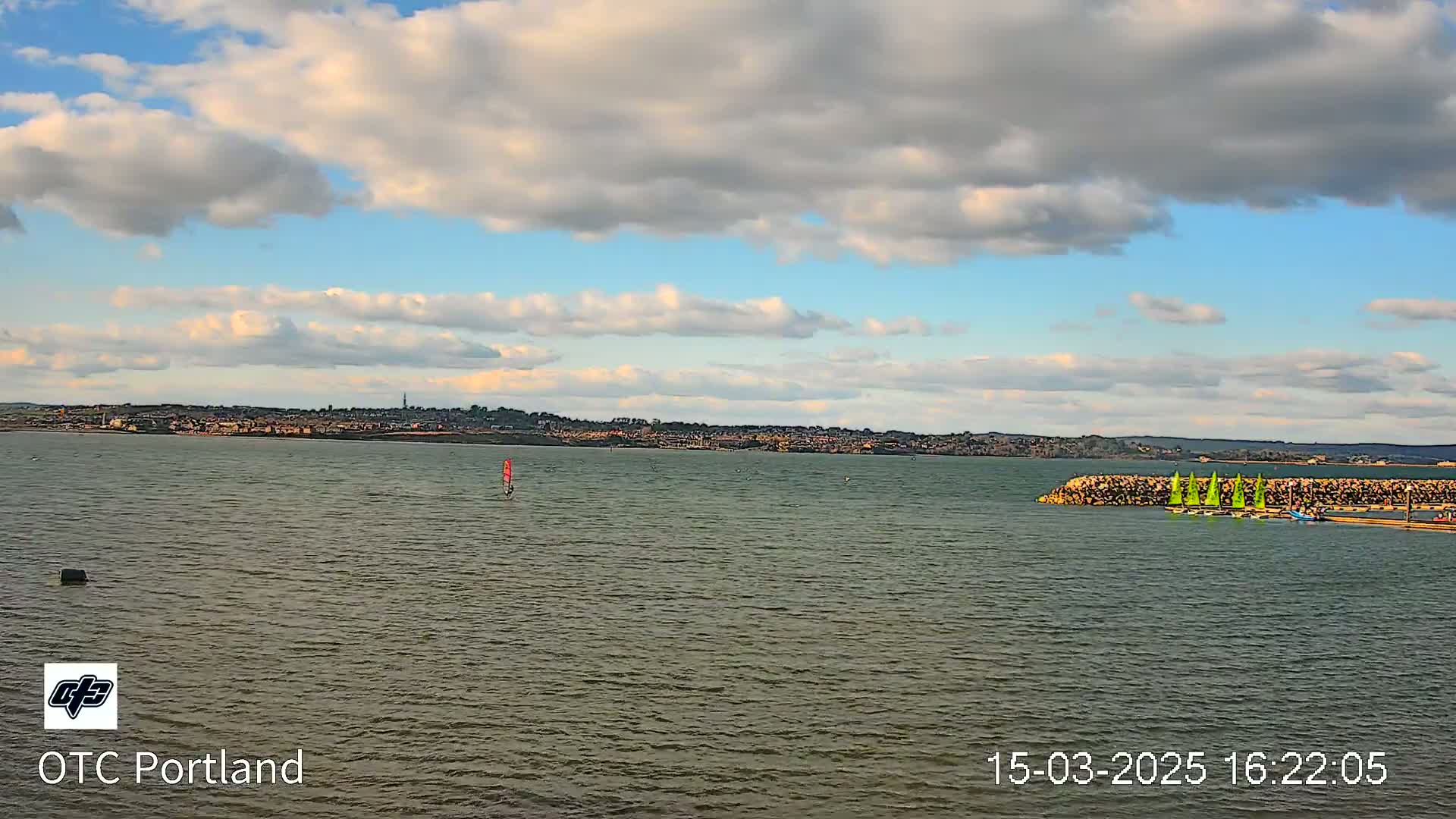 A partly cloudy sky overlooks a calm body of water with several sailboats near a rock jetty and a distant town visible on the horizon.