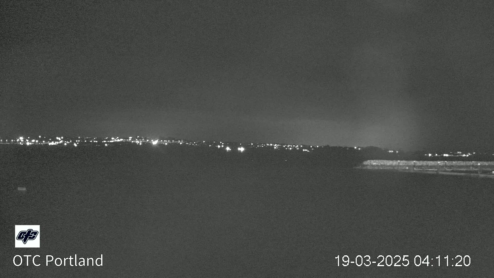 A nighttime, low-light view shows a distant line of city lights across a dark, flat landscape under a cloudy sky.
