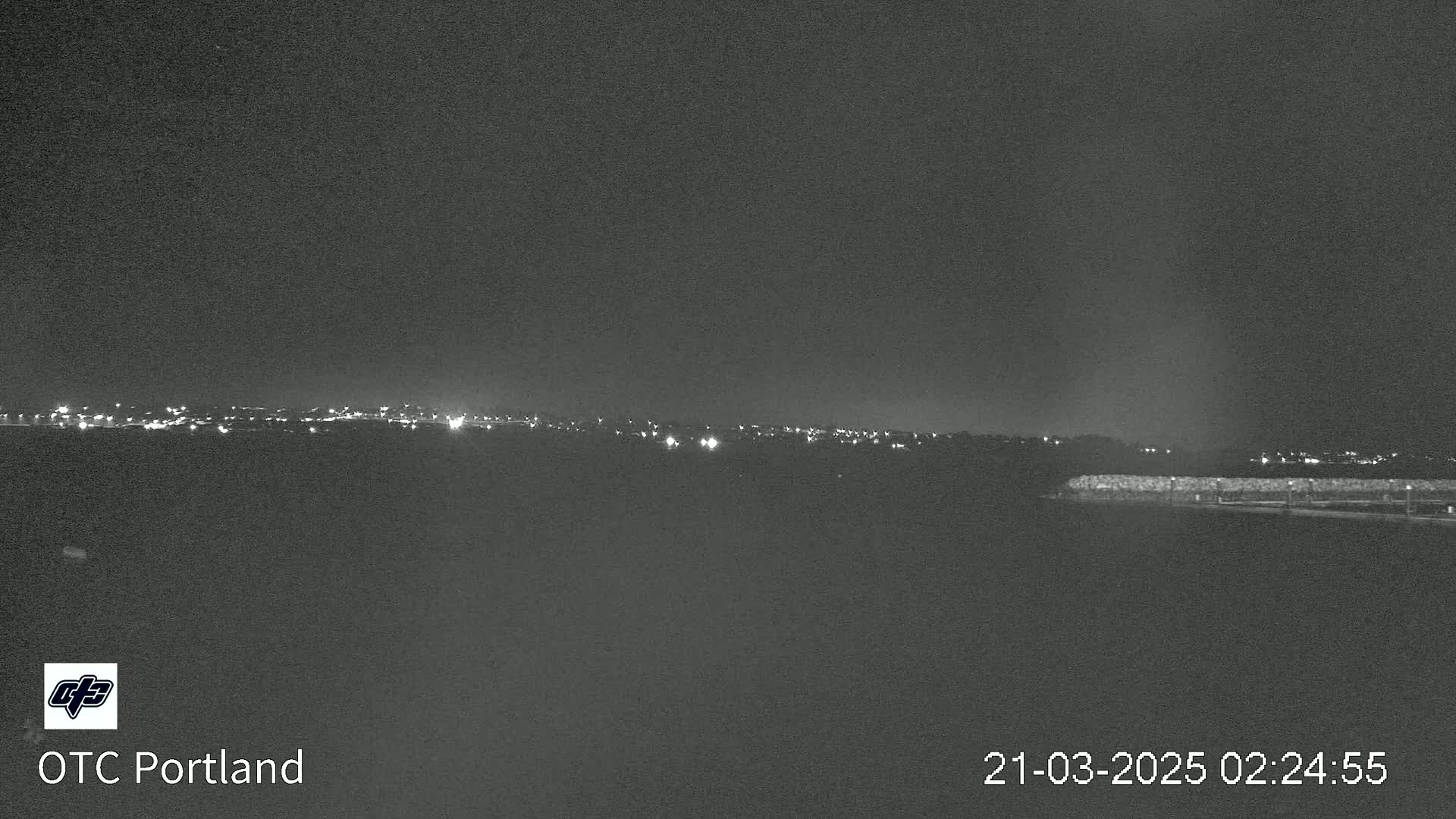 A nighttime long shot shows a distant line of city lights across a dark landscape, with a dark structure in the foreground under a clear, dark sky.