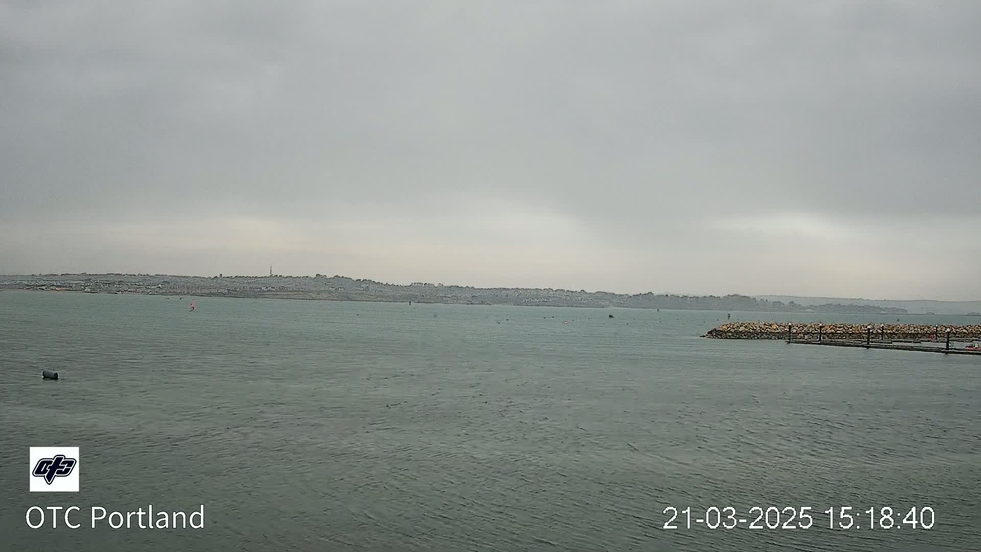 A calm body of water stretches toward a distant, overcast shoreline under a gray sky.