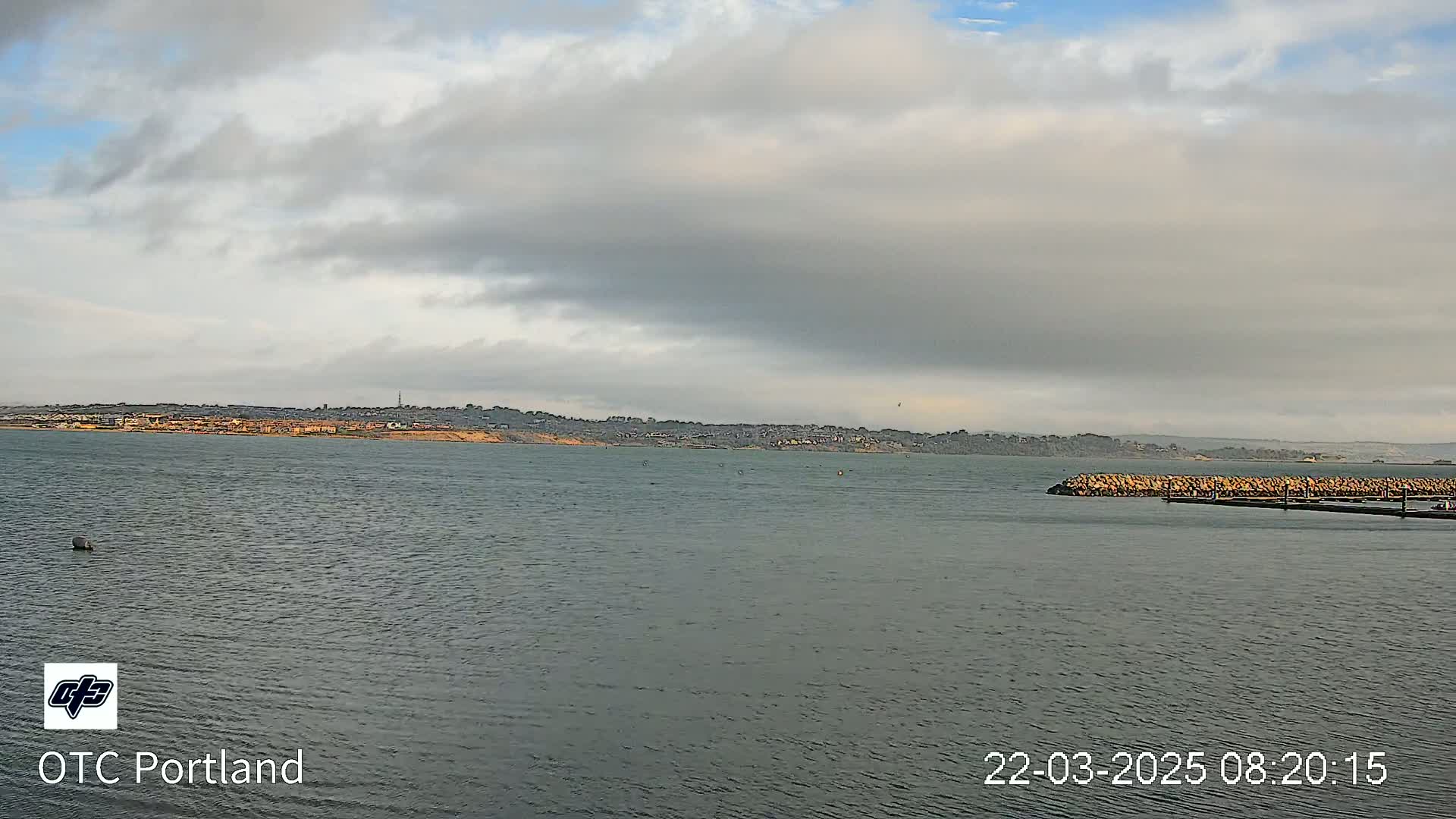 A body of calm water stretches out to a distant shoreline under a mostly cloudy sky.
