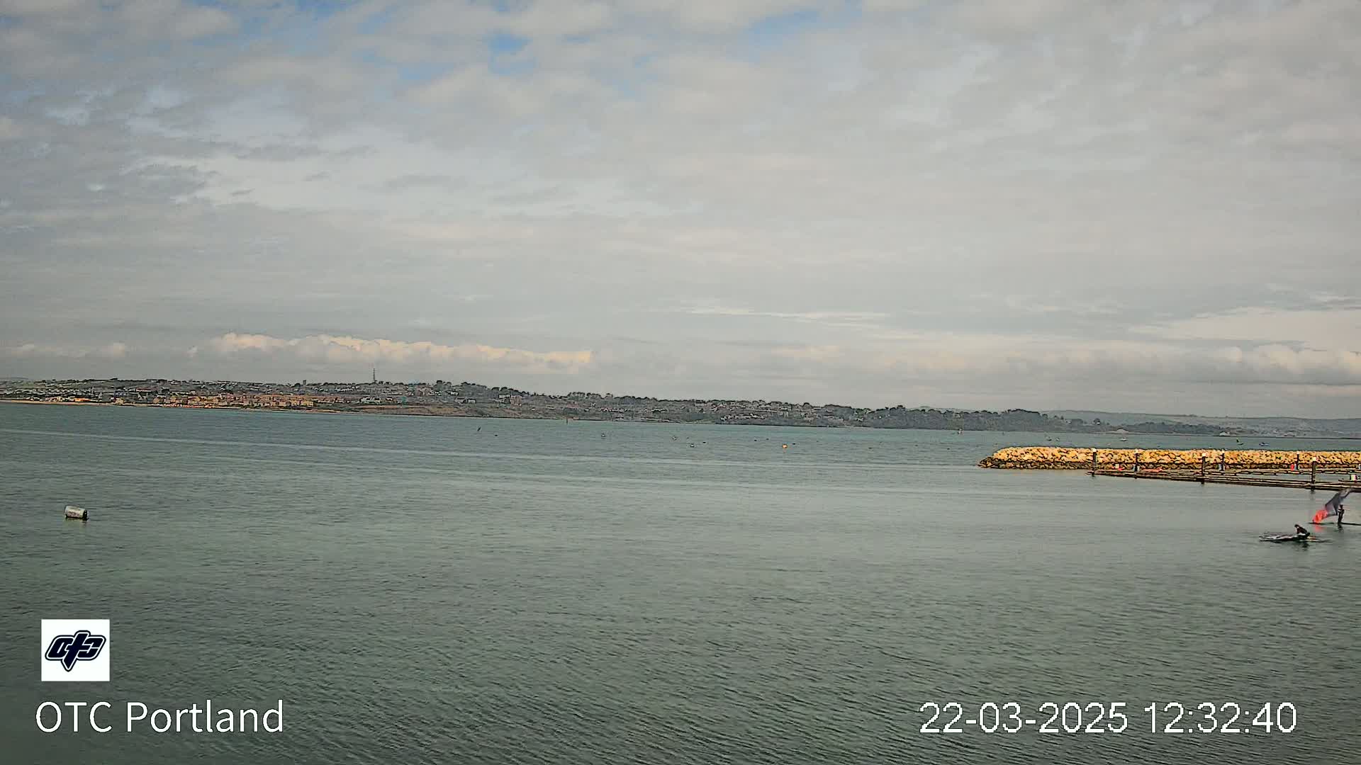 A calm body of water with a distant shoreline under a cloudy sky shows a person windsurfing near a pier.
