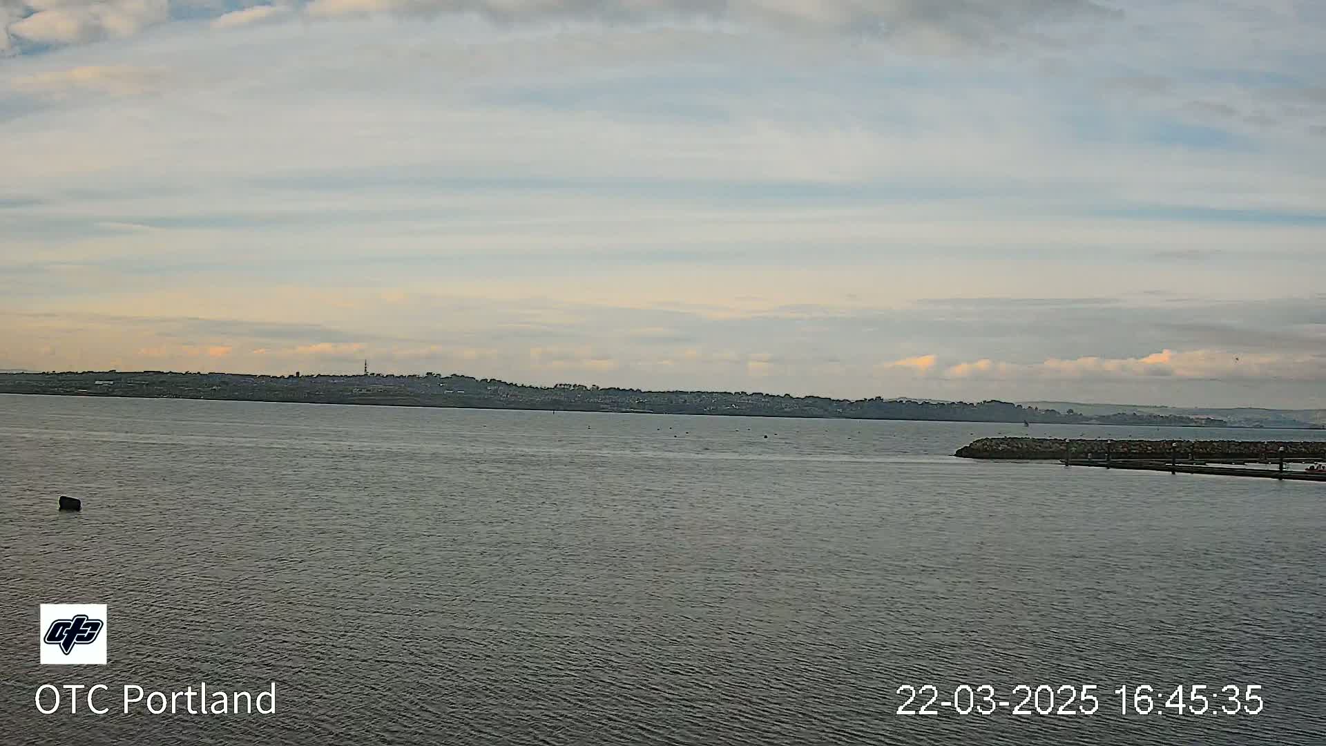 A calm body of water shows a distant shoreline under a mostly cloudy sky.