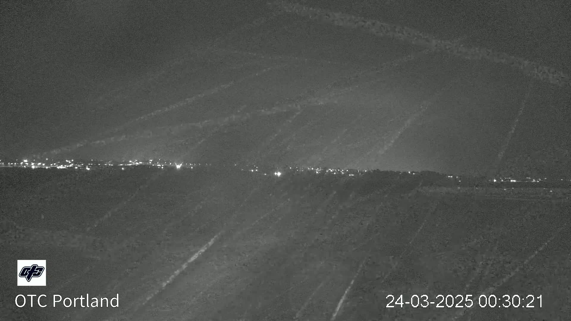 A nighttime, low-light view shows a distant city skyline illuminated with scattered lights, under a dark sky with streaks possibly caused by rain or snow.