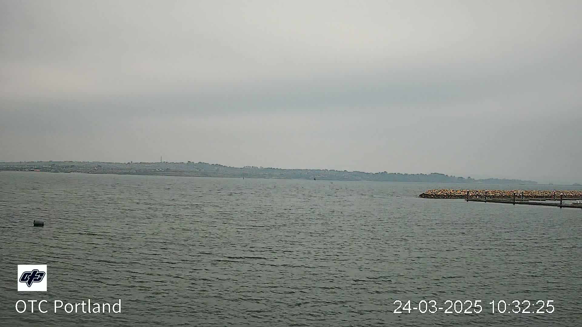 A body of water with a distant shoreline under an overcast sky shows a small rock jetty in the foreground.