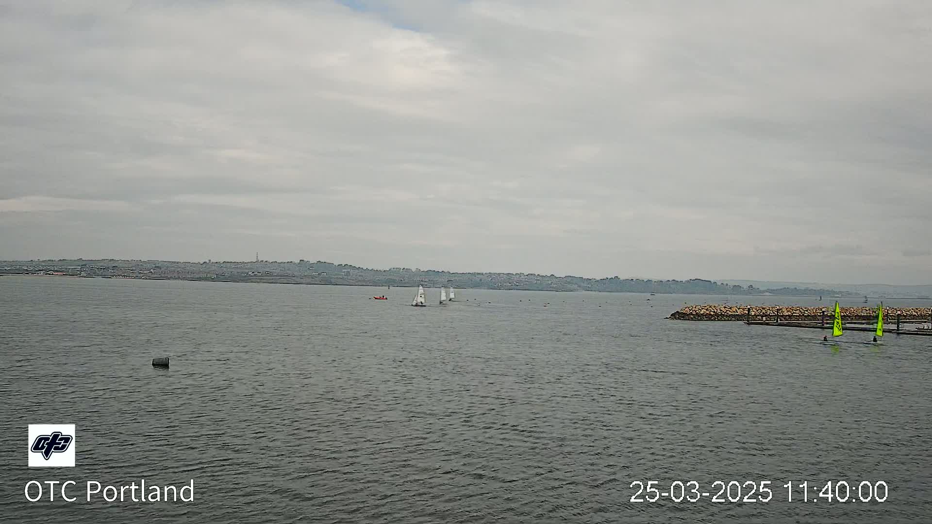 Several sailboats are visible on a gray, overcast day on a body of water with a distant shoreline.