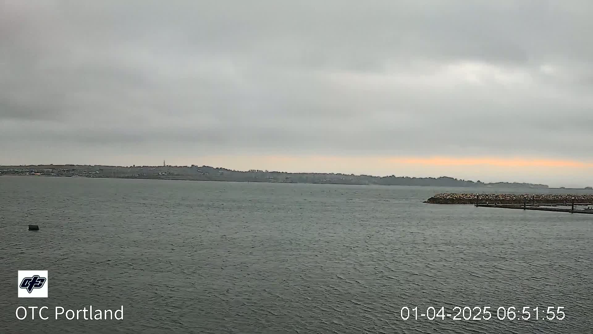 A calm body of water is shown under an overcast sky with a faint orange hue on the horizon, and a distant shoreline is visible.