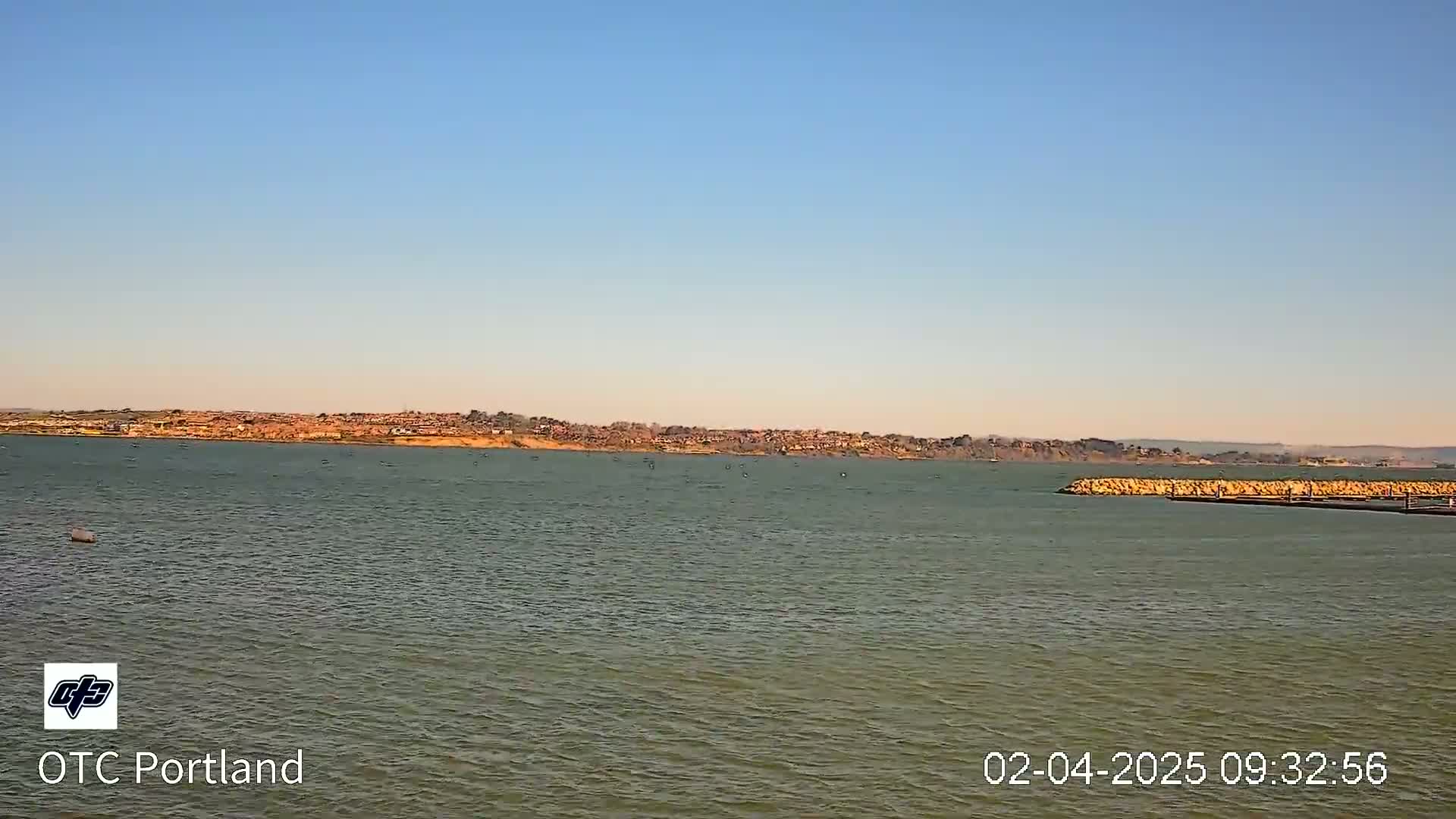 A calm body of water shows a distant shoreline with buildings under a clear, bright sky.