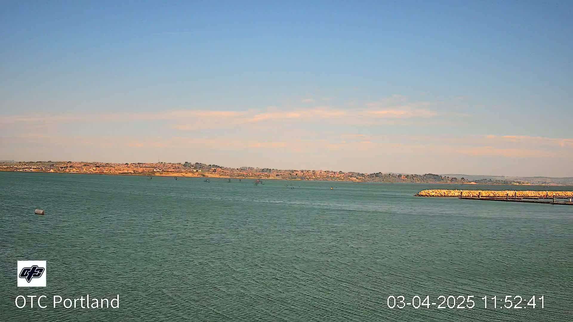 A calm, greenish-blue body of water stretches toward a distant shoreline dotted with buildings under a mostly clear, light-blue sky.