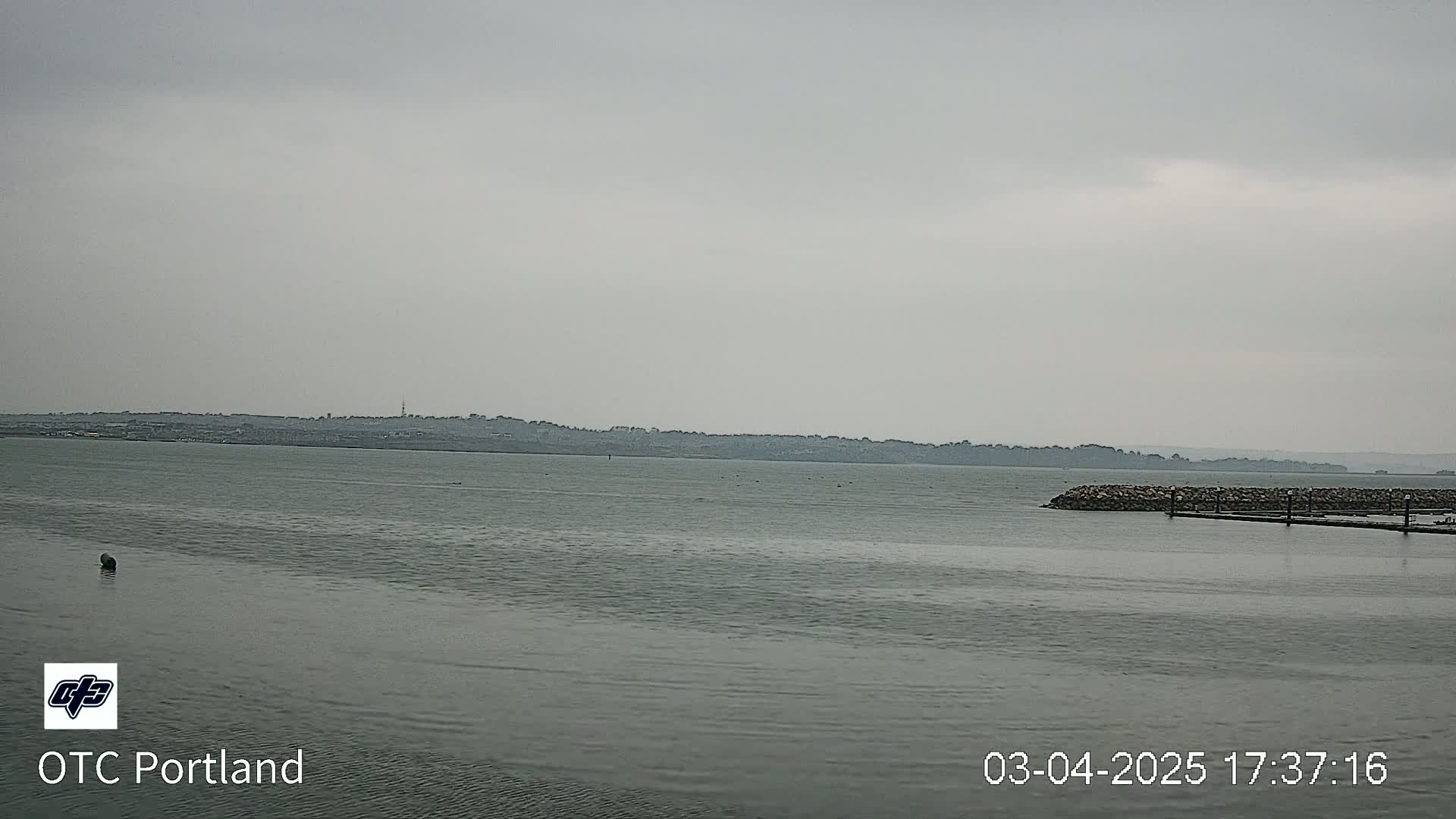 A calm body of water under an overcast sky shows a distant shoreline with low-lying buildings.