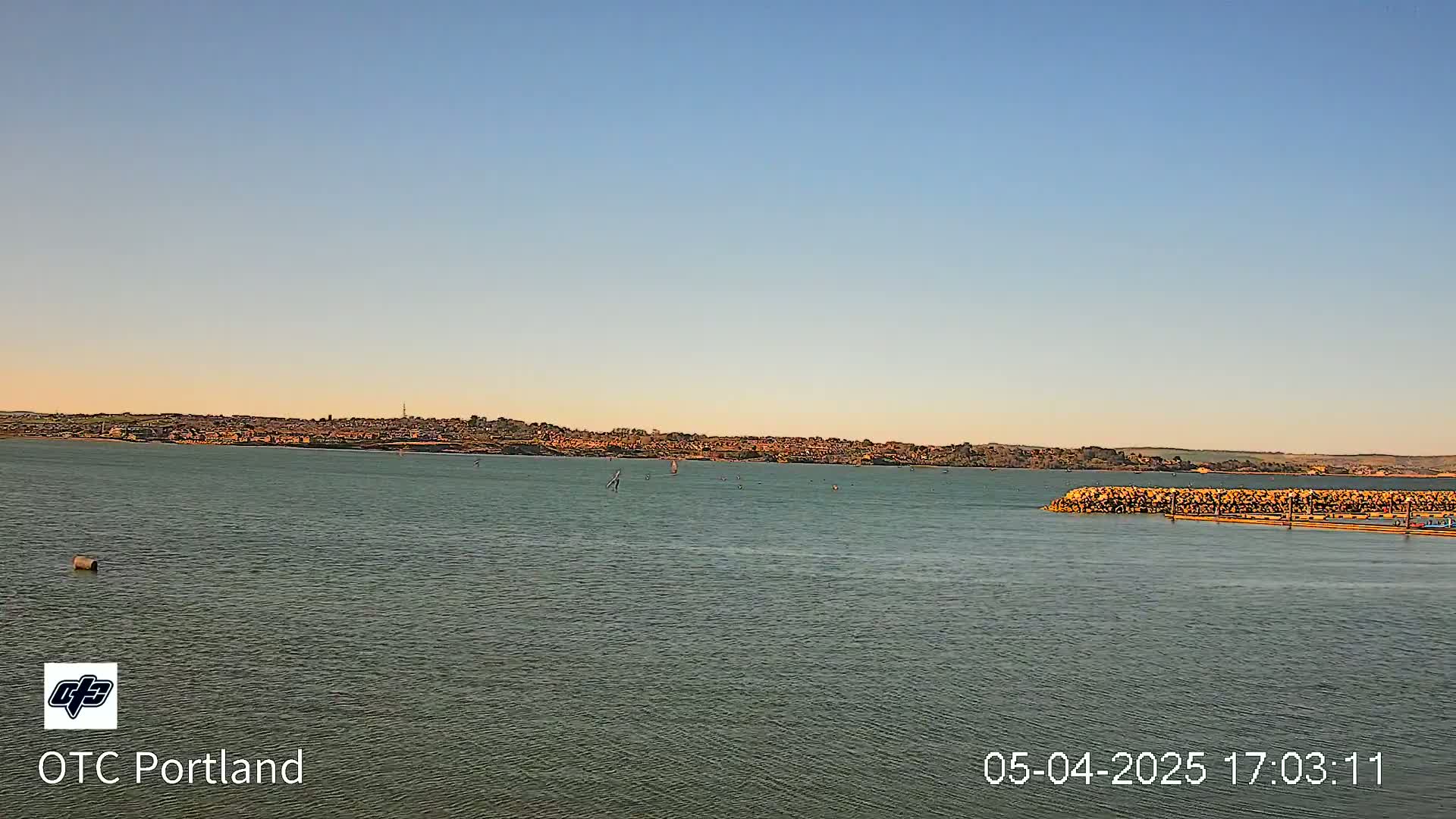 A calm body of water shows several sailboats under a clear, pale-blue sky at sunset, with a distant town visible on the horizon.