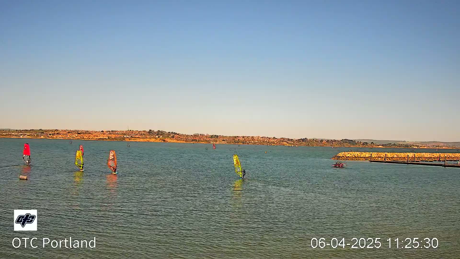Several windsurfers are sailing on a calm body of water under a clear, sunny sky, with a distant shoreline visible in the background.