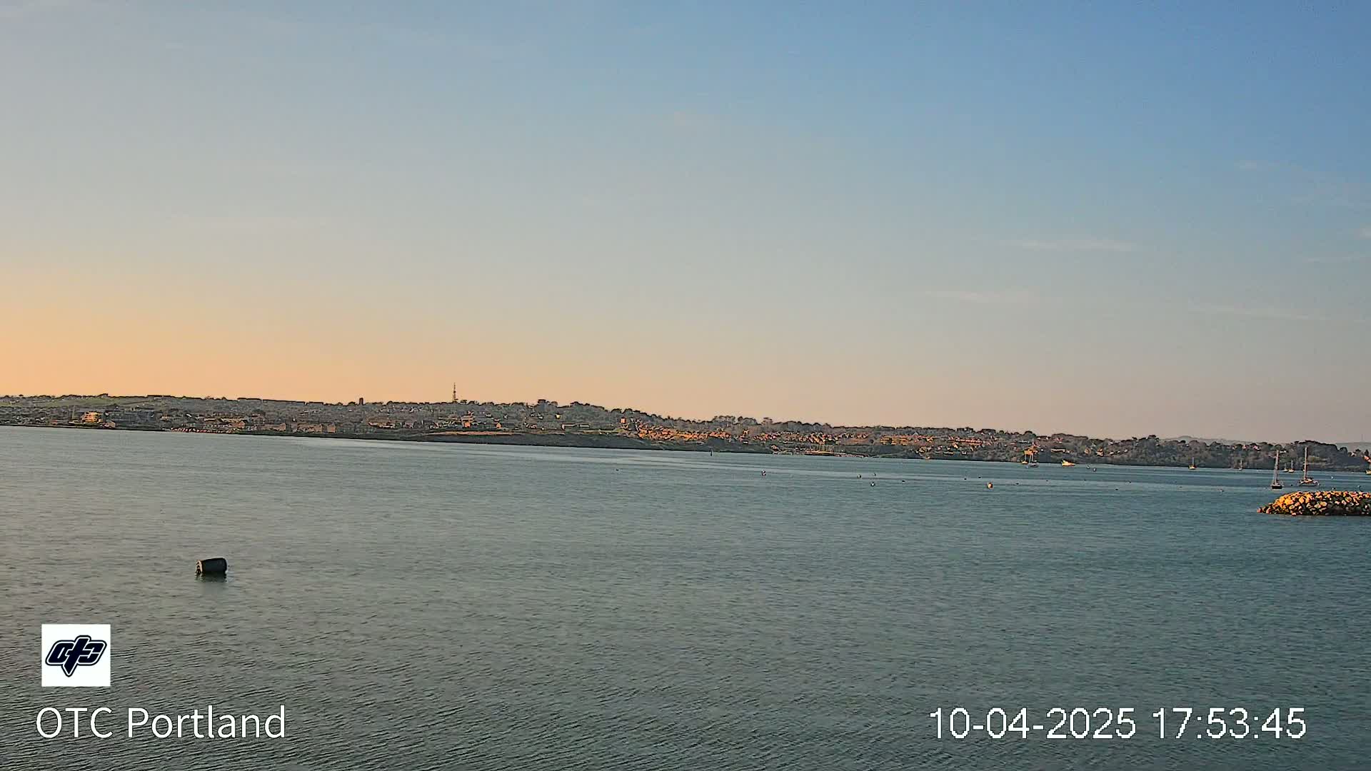A calm body of water stretches towards a distant town under a clear, pale-orange sunset sky.