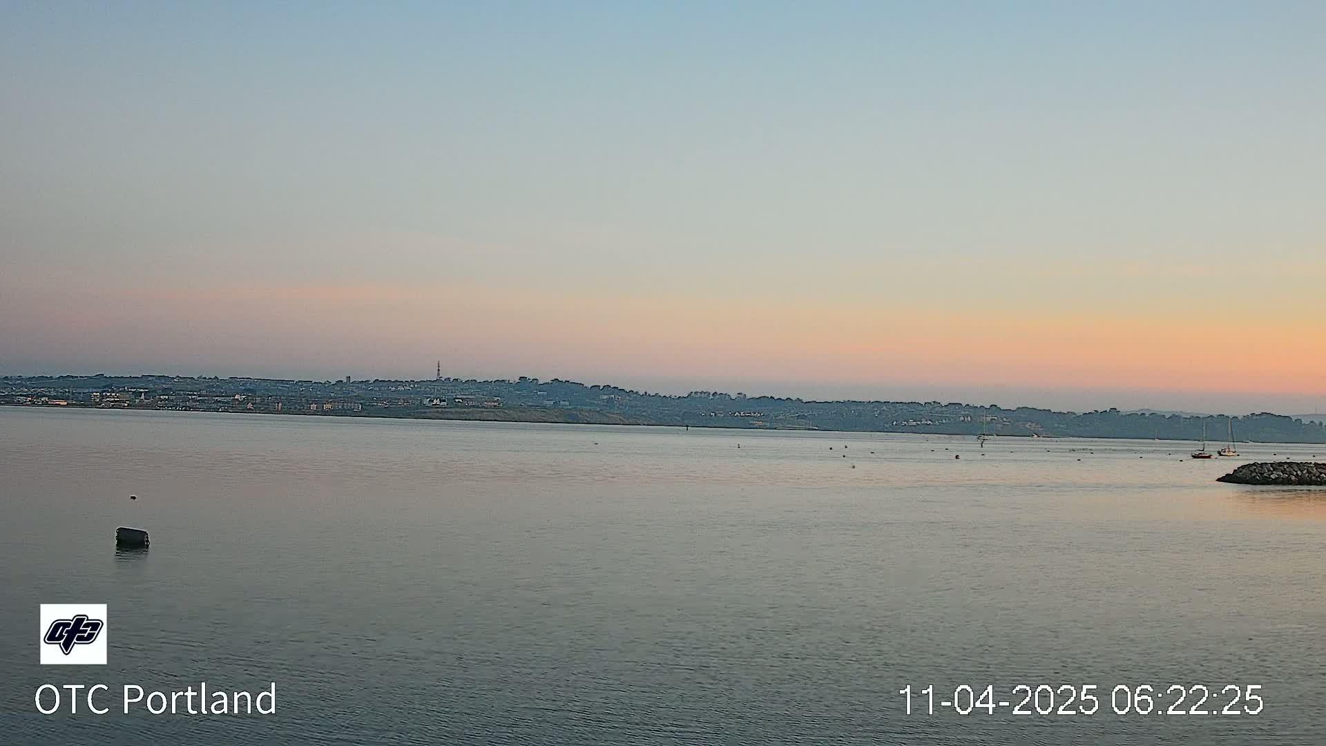 A calm body of water at sunset shows a distant cityscape under a pale pink and orange sky.
