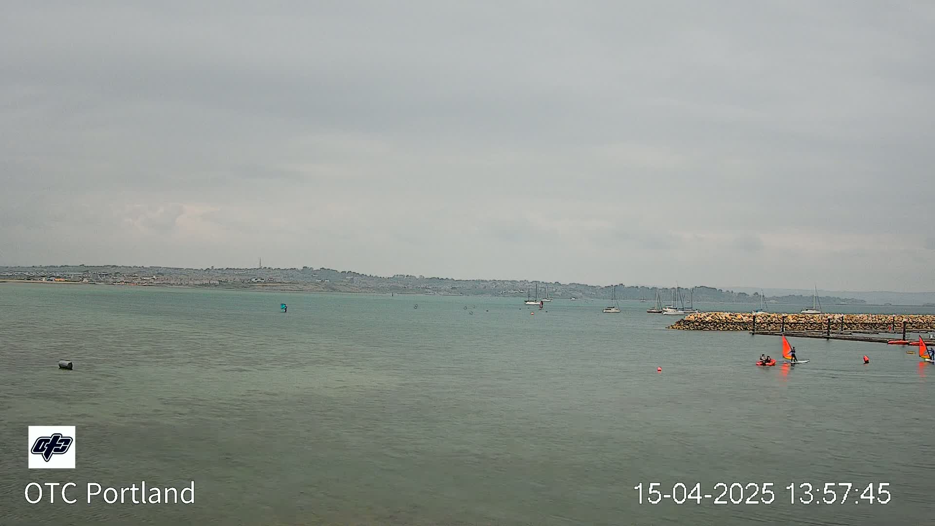 A calm body of water under an overcast sky shows several sailboats and small watercraft near a stone breakwater with a distant shoreline.