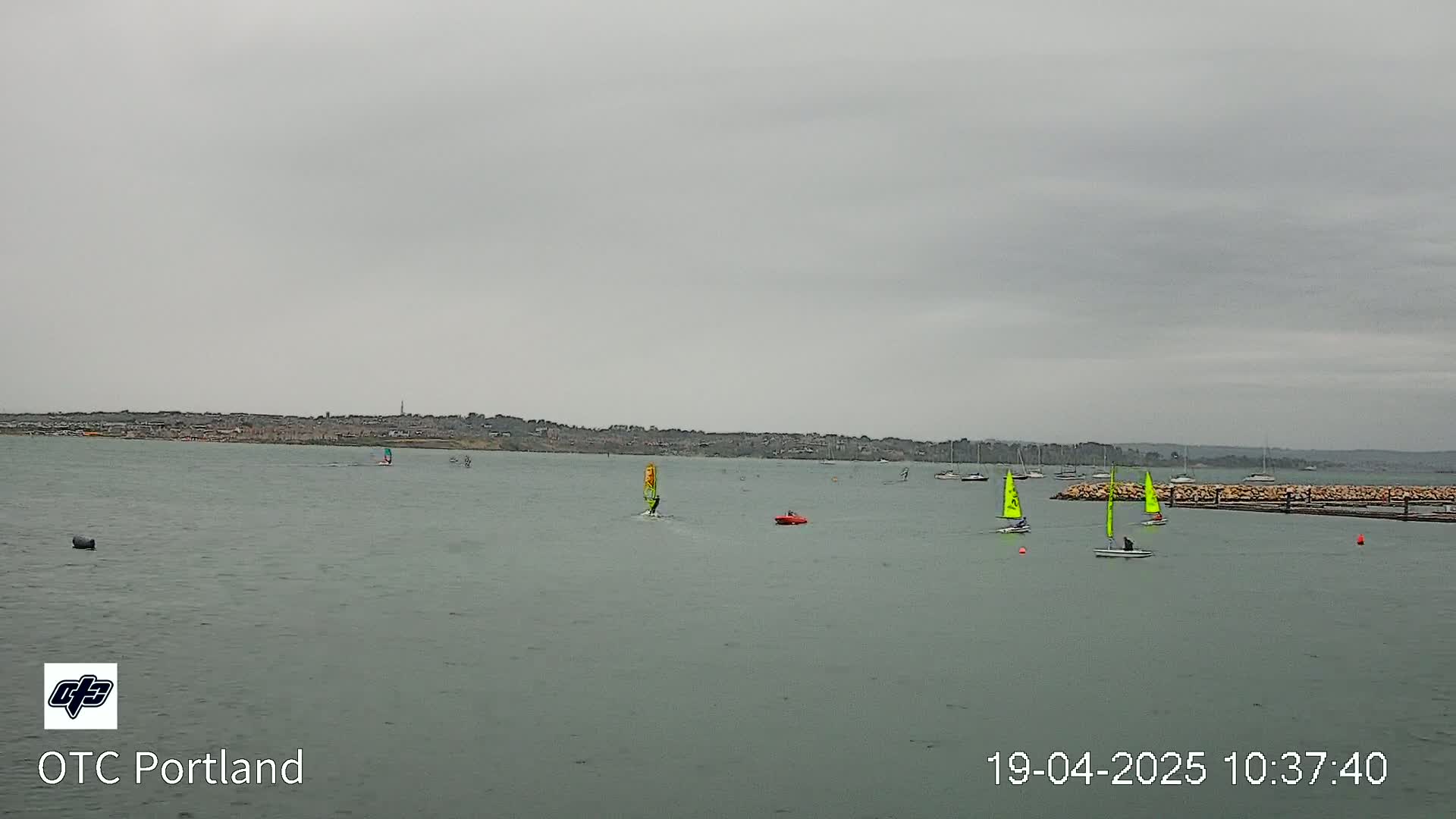Several sailboats and windsurfers are on a calm, gray body of water under an overcast sky.