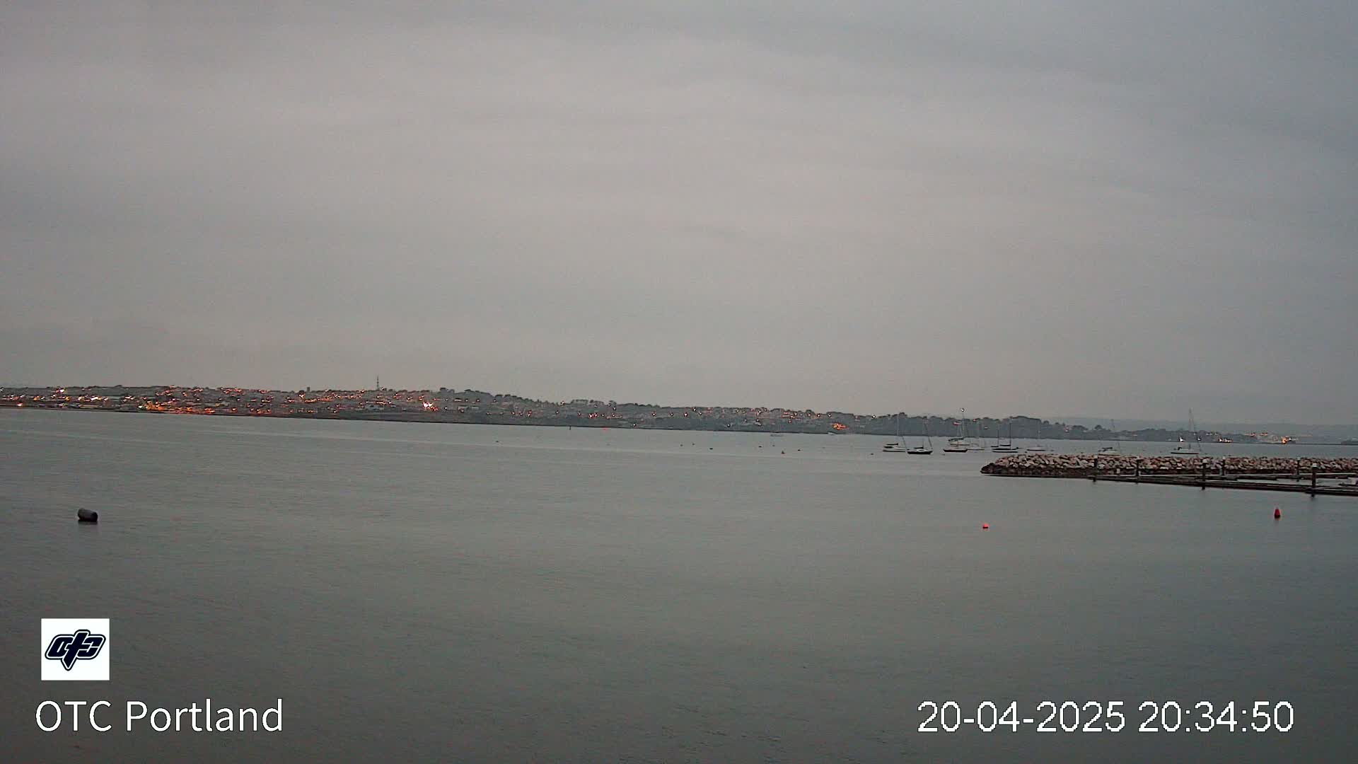 A calm body of water shows a distant city skyline under an overcast sky, with several boats moored near a rocky jetty.