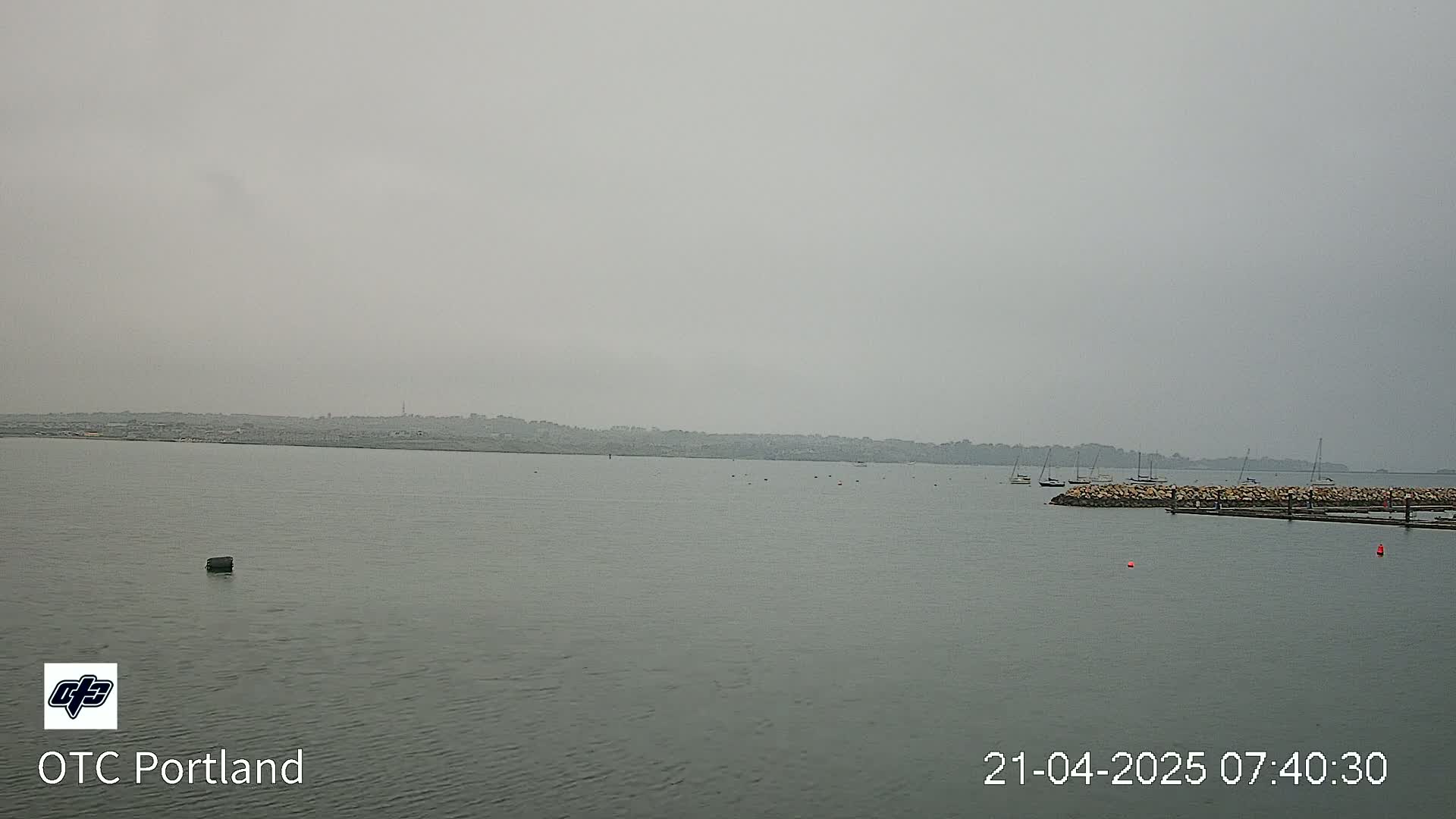 A hazy, gray day shows a calm body of water with several sailboats moored near a rock jetty, and a distant shoreline visible in the background.