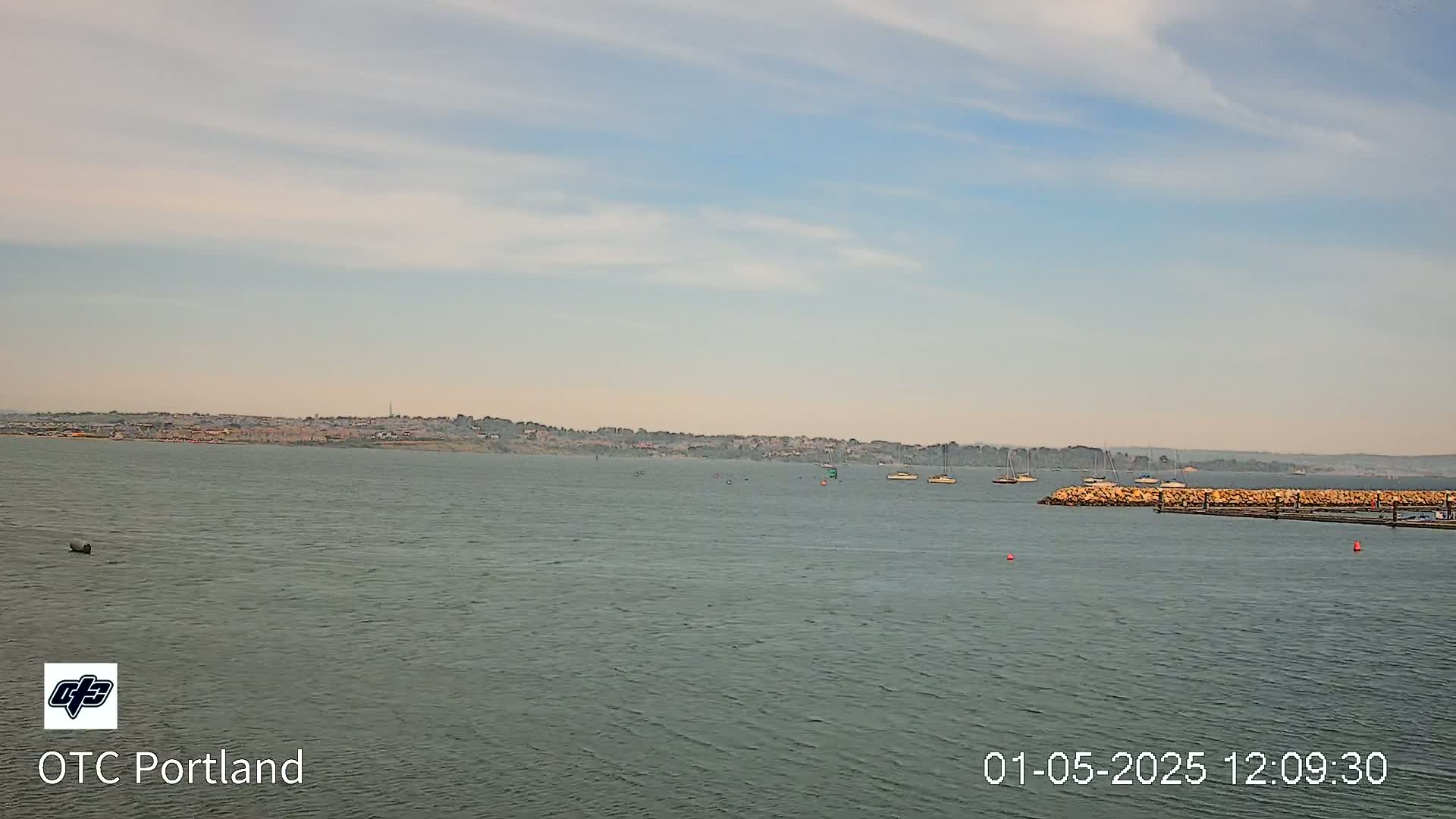 A calm body of water shows several sailboats moored near a low stone breakwater with a distant shoreline under a mostly clear, light blue sky.