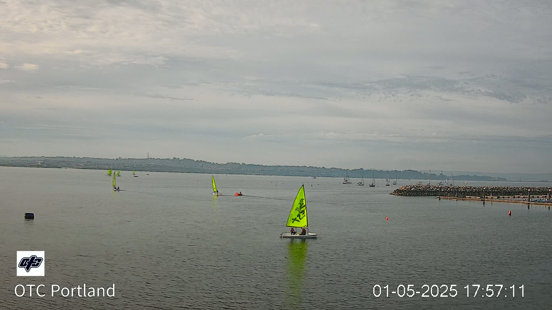 Several sailboats are on a calm body of water under an overcast sky, with a distant shoreline visible.