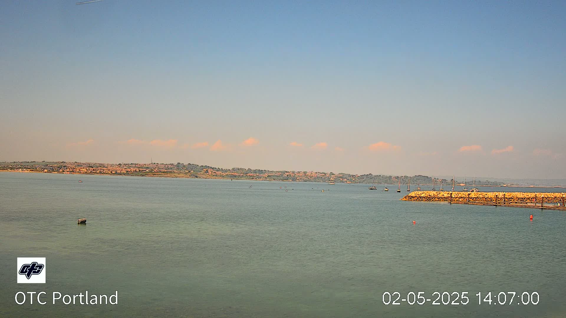 A calm body of water shows a distant shoreline with buildings under a mostly clear sky with a few small clouds.