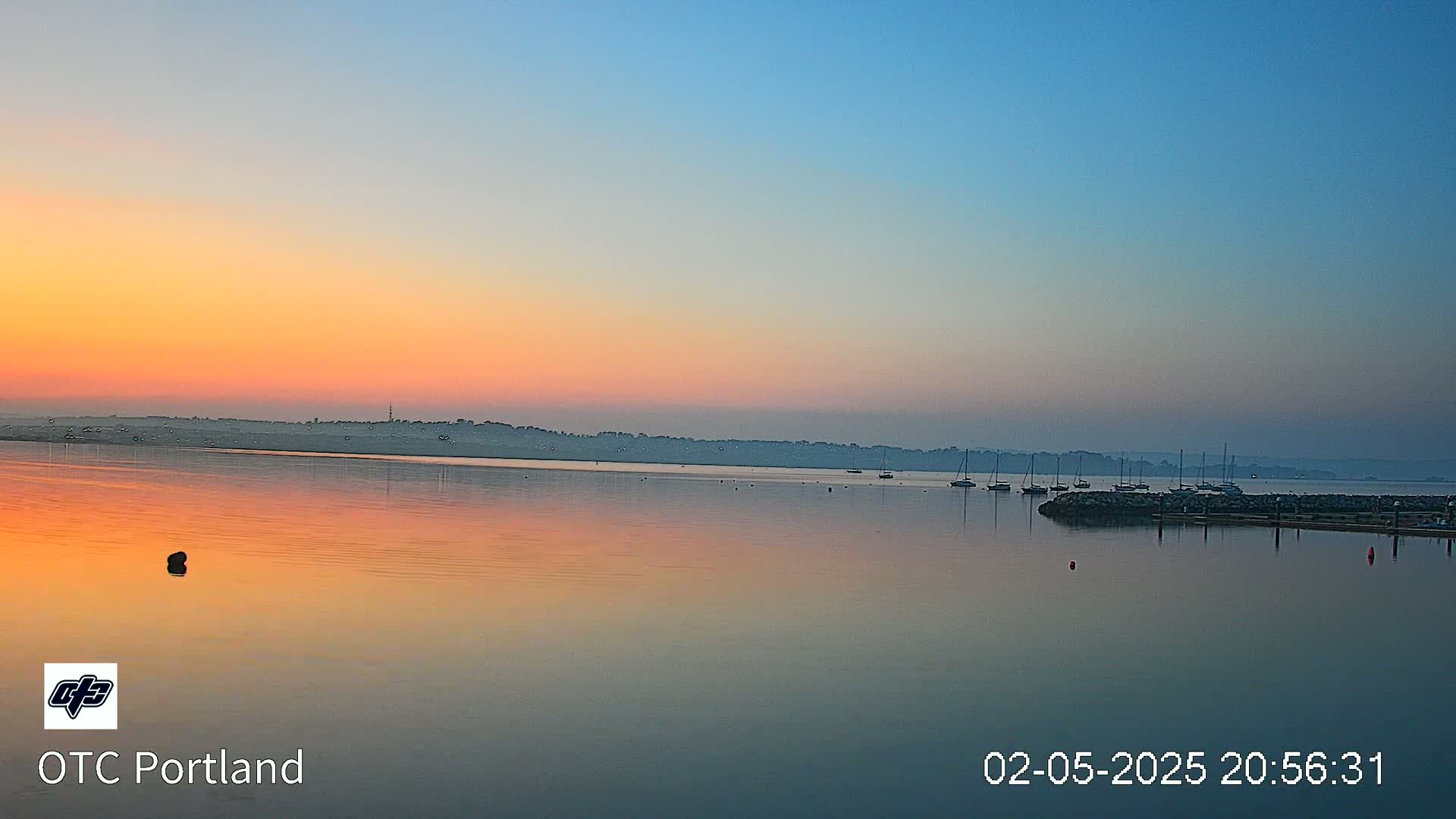 A calm body of water reflects a pastel sunrise sky, with several sailboats moored in the distance.
