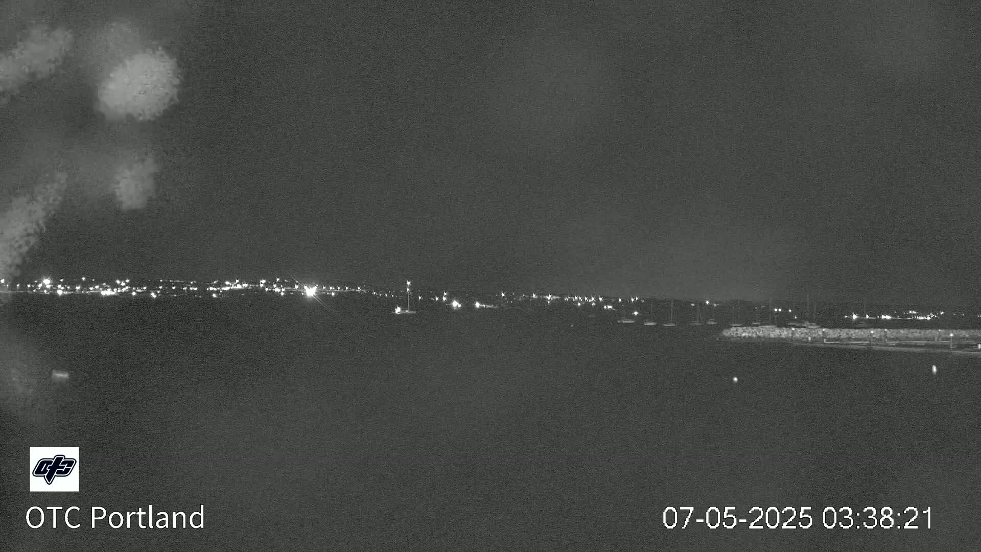 A nighttime long shot shows a dark body of water with a distant shoreline of lights, a low stone wall, and a few boats.