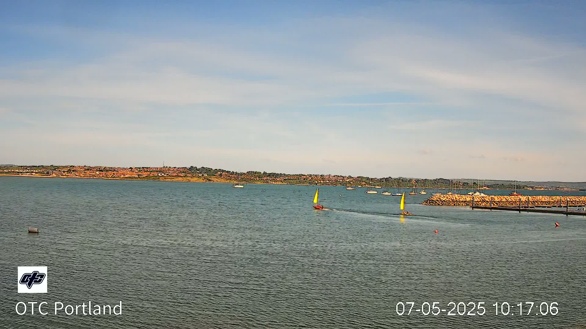 Under a mostly clear sky, a body of water shows several sailboats and a breakwater with docked boats near a distant shoreline of buildings.