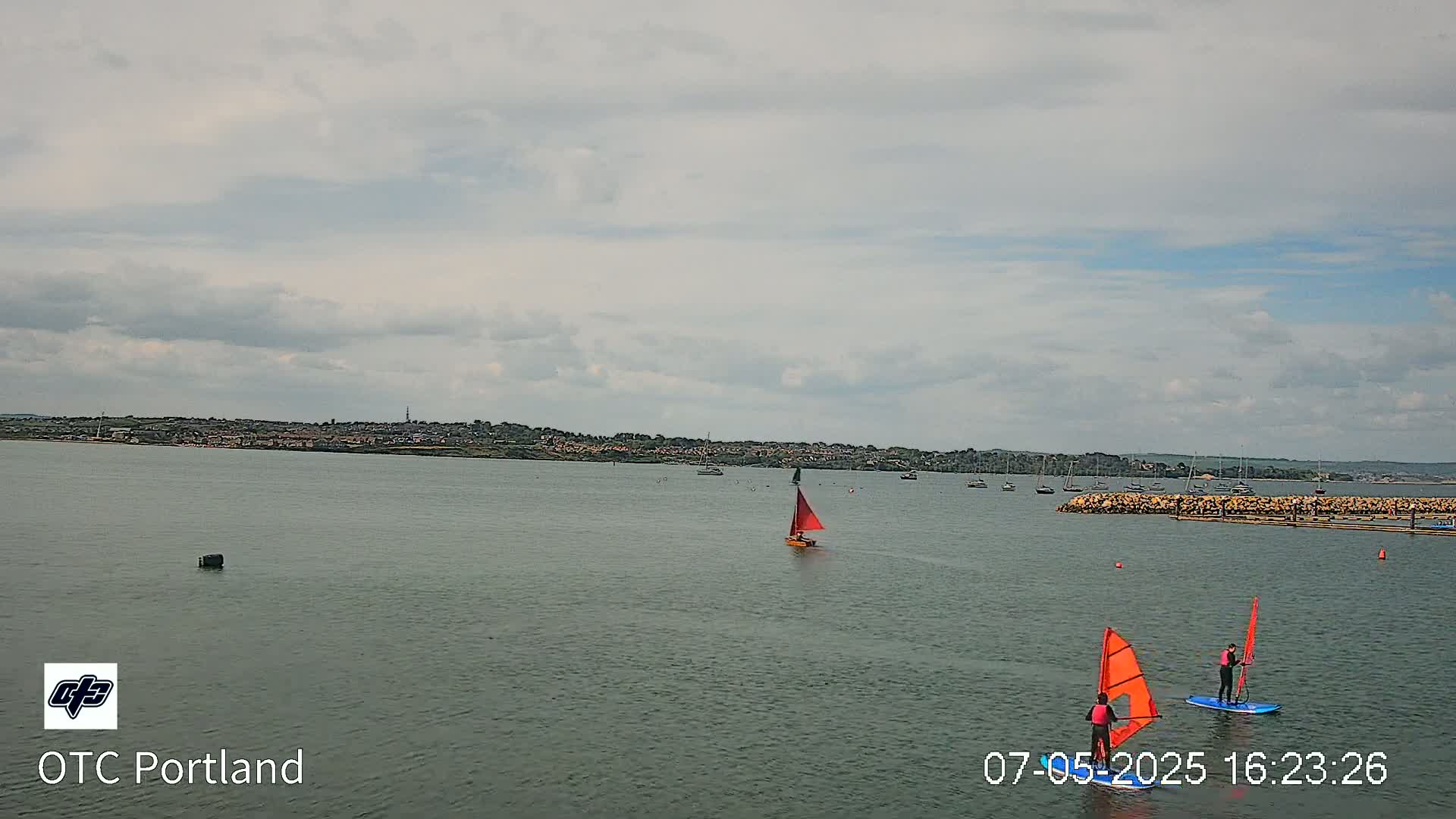 A calm body of water shows a small sailboat and two windsurfers under a mostly cloudy sky, with a distant shoreline visible in the background.